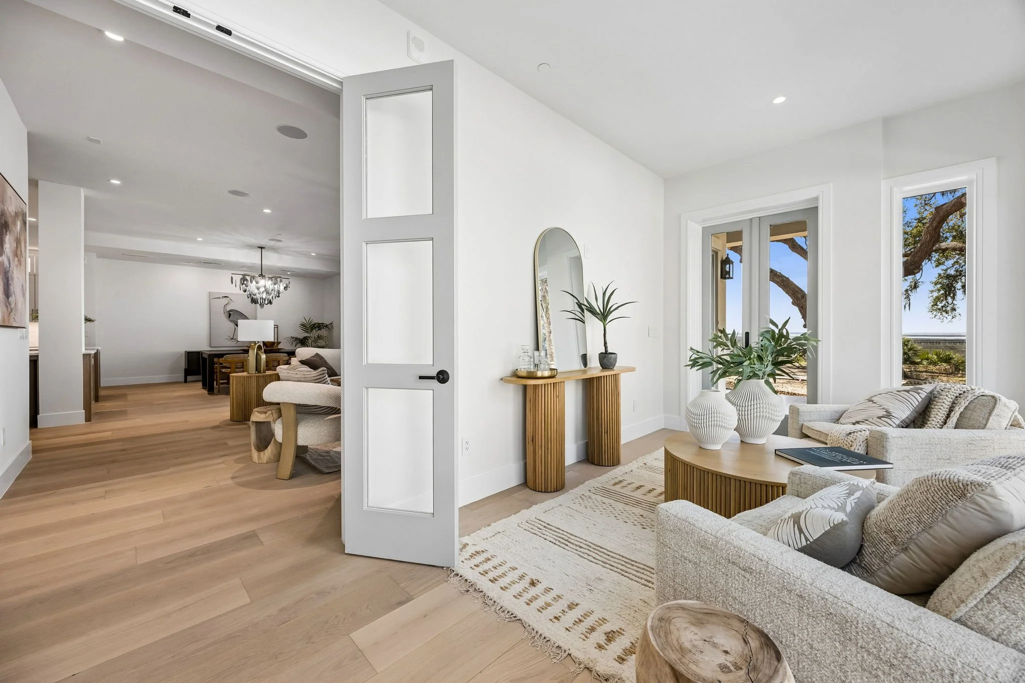 Bright living room with white walls and large windows showing outdoor trees, fitted with beige sofas, a wooden coffee table, white decorative vases, and greenery, with light wooden flooring and modern decor.