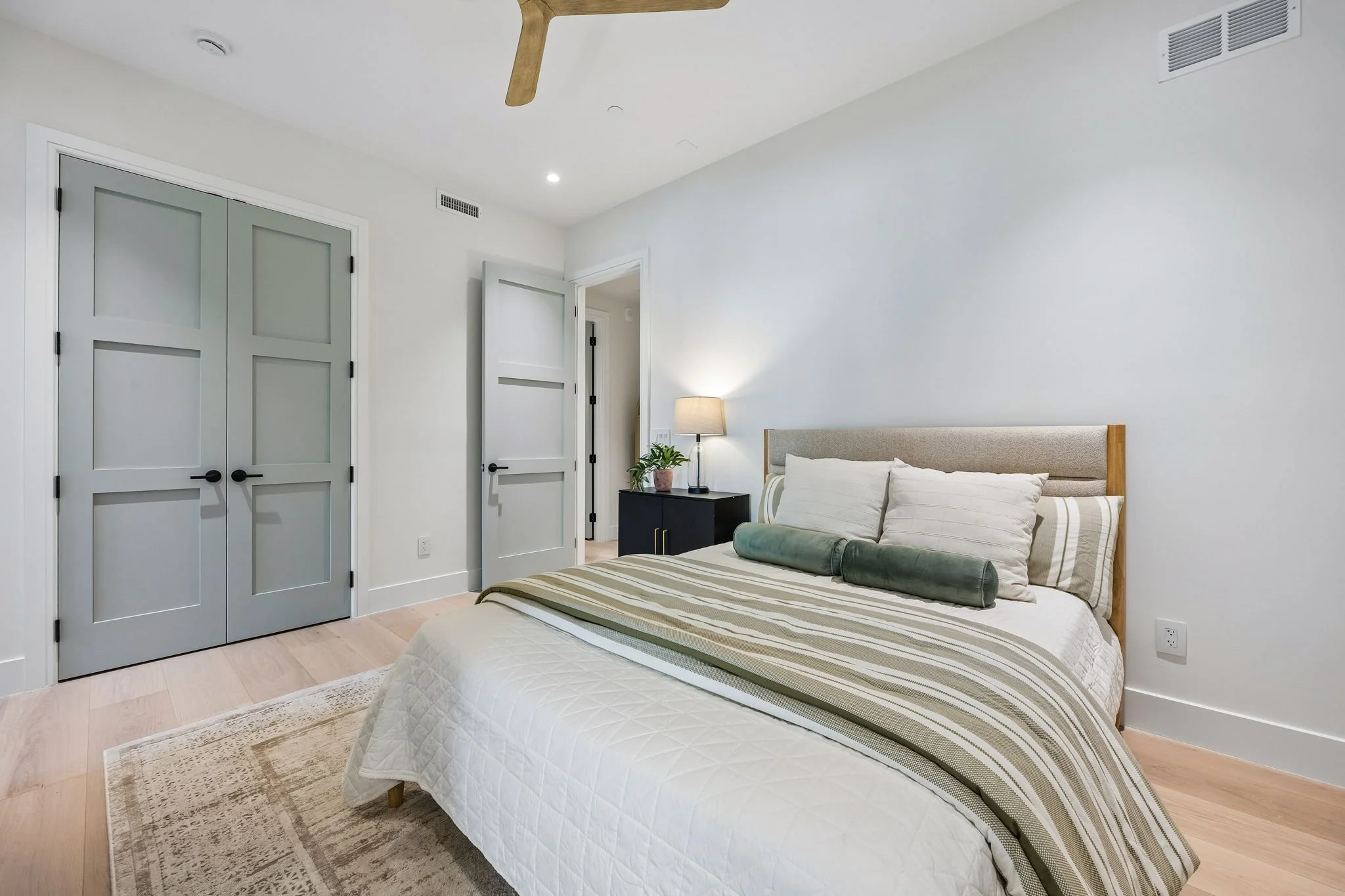 A bedroom with a bed, striped bedding, and pillows, near a black nightstand with a lamp and plant. Light-colored walls, wooden flooring, and closed double doors.