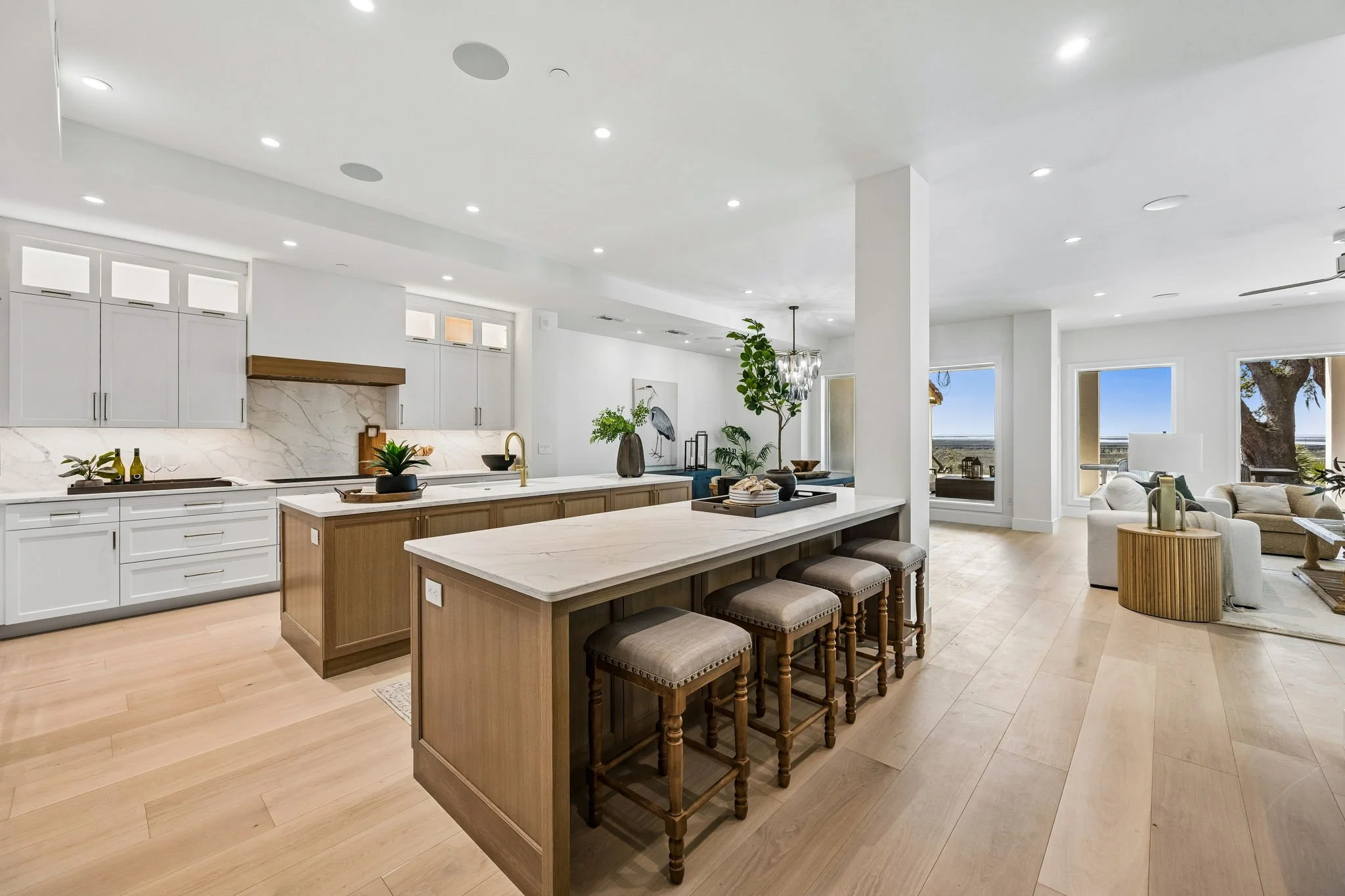 Open-concept kitchen and living room with white walls, light wood floors, and large windows showing outdoor scenery. The kitchen features white cabinets, a marble backsplash, a wooden kitchen island with a marble countertop, and barstools. The living