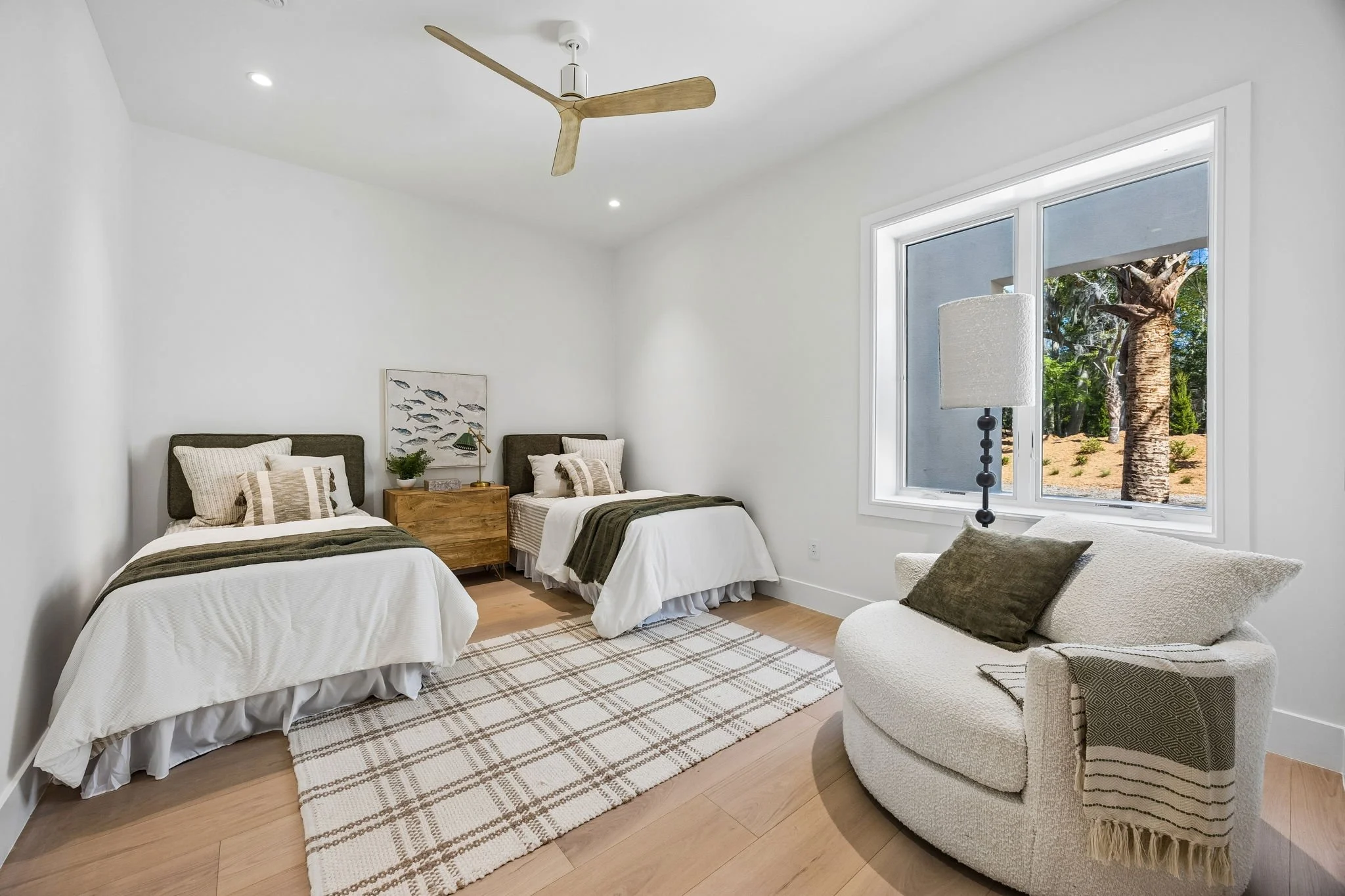 A bright bedroom with white walls, two twin beds with gray headboards and striped bedding, a small wooden nightstand with a lamp and potted plant between the beds, a white round plush chair with pillows near a large window showing outdoor trees, a pa