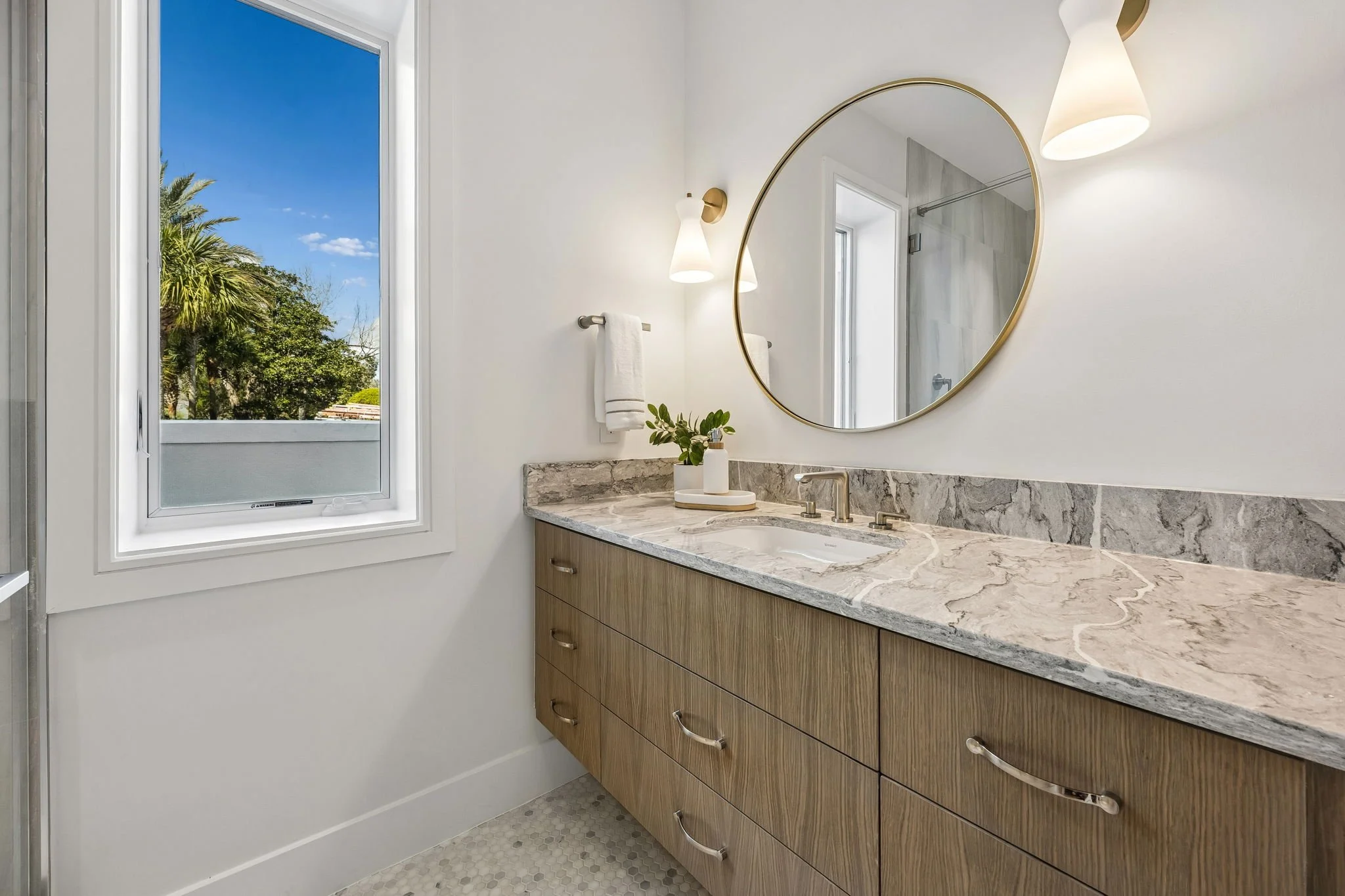 Bathroom vanity with marble countertop, round mirror, white walls, window showing greenery outside, towel rack with a white towel, and modern lighting fixtures.