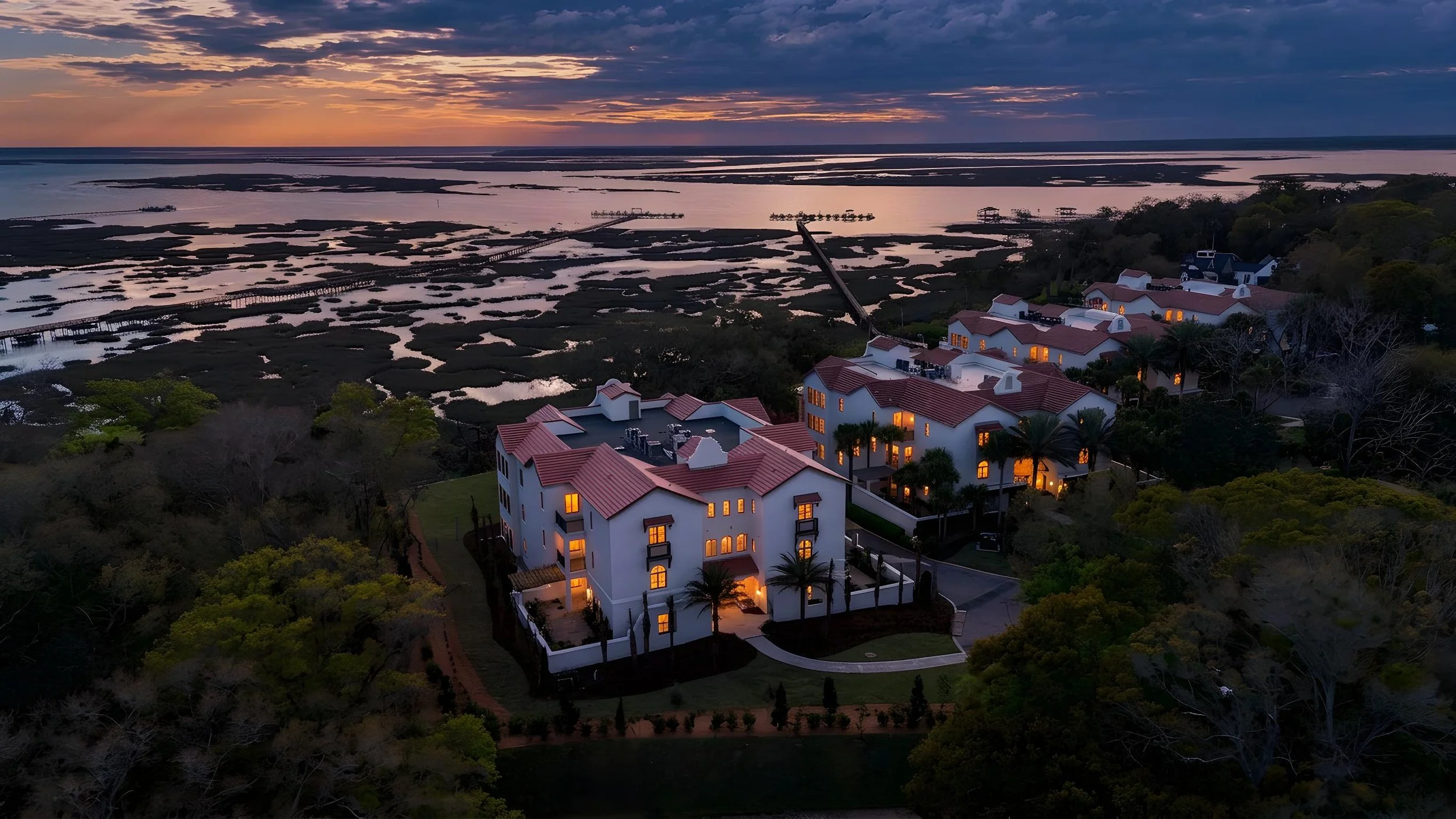 The Landings On Amelia River. An aerial view of a coastal residential complex with white buildings, red roofs, and landscaped gardens during sunset, overlooking marshlands and water channels.