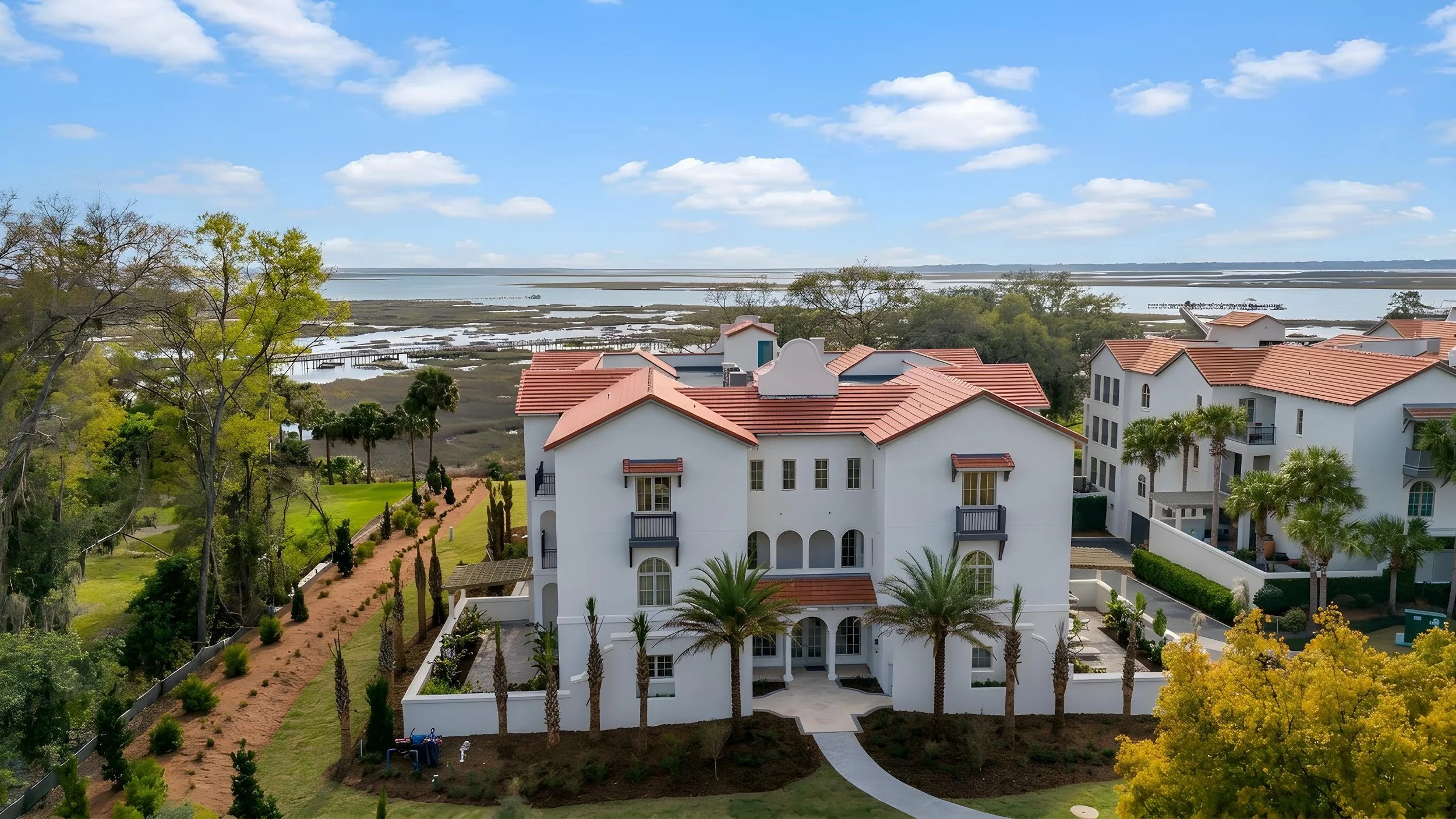 The Landings On Amelia River. A large white multi-story house with a red tile roof, surrounded by palm trees and other greenery, with water and marshlands in the background under a partly cloudy sky.