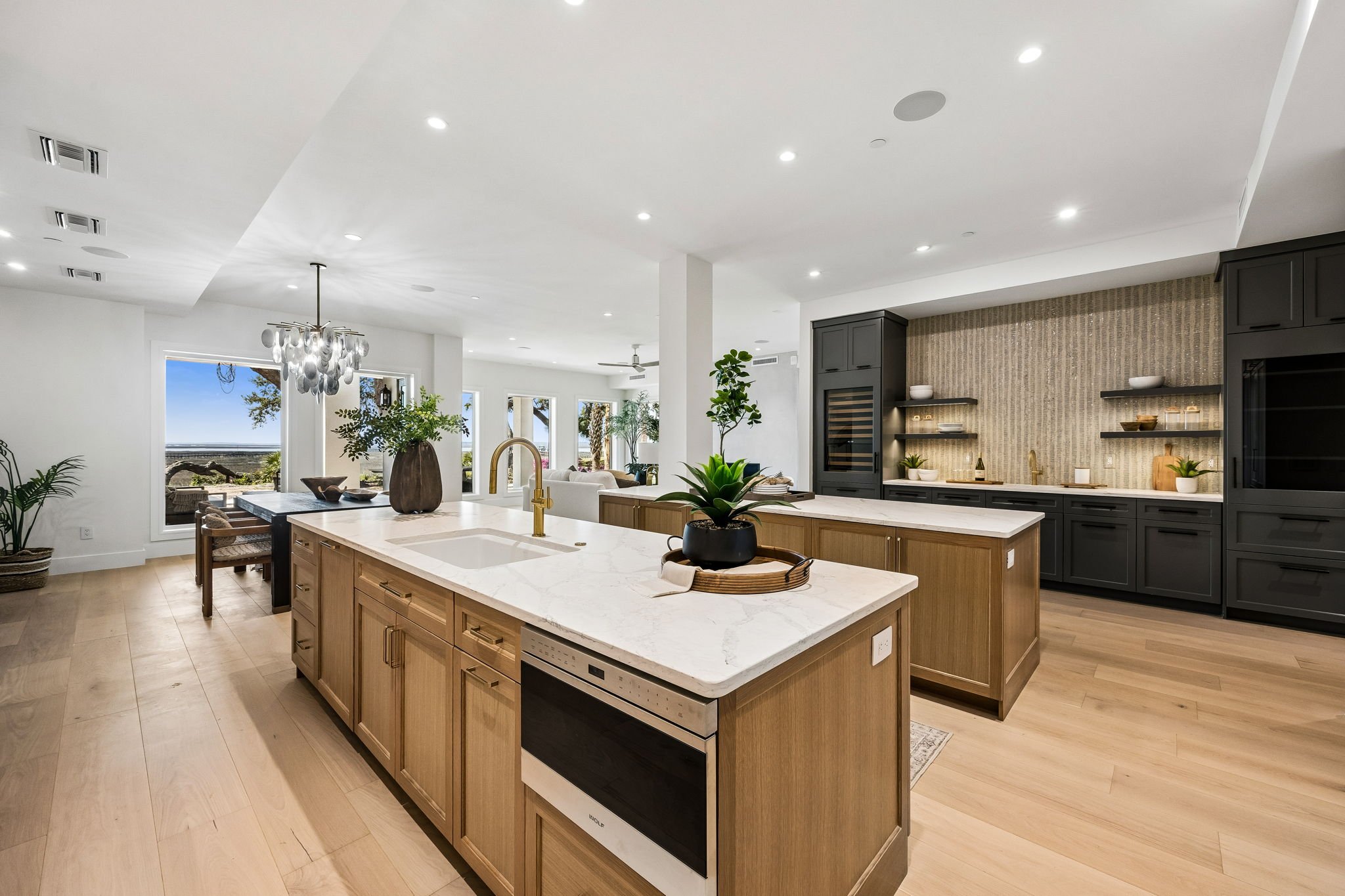 Modern kitchen with light wood cabinets, a white marble island, black upper cabinets, and a sitting area with trees visible through large windows.
