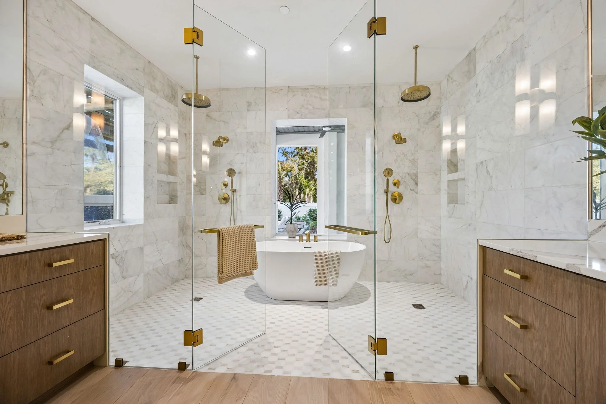 Modern bathroom with marble walls, glass shower doors, a freestanding bathtub with a window behind it, wooden vanities with gold handles, and gold fixtures including showerheads and towel bars.