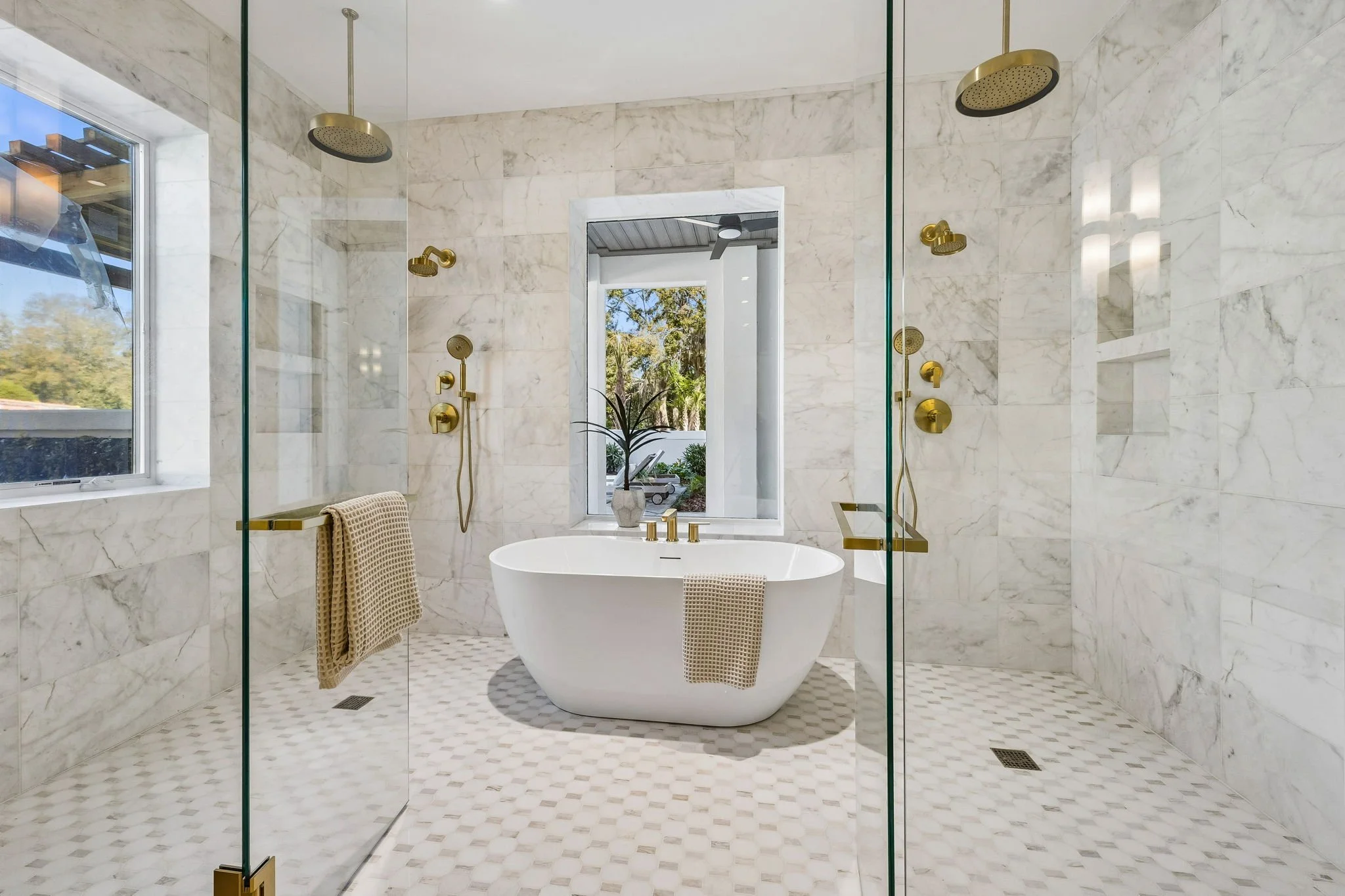 Modern bathroom with marble walls and floor, a freestanding bathtub, and a double shower with gold fixtures. A window with a view of trees outside and a potted plant on the windowsill.