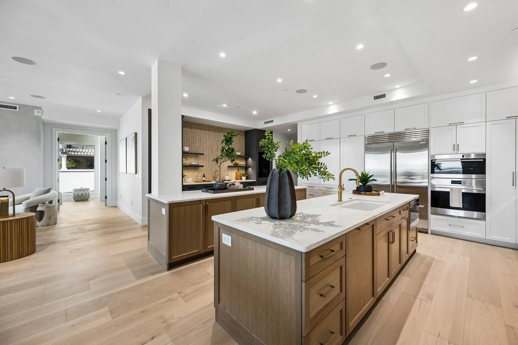 Modern open-concept kitchen with white and wood cabinetry, a large kitchen island with a white countertop, and stainless steel appliances. Decorated with green plants and black vases.