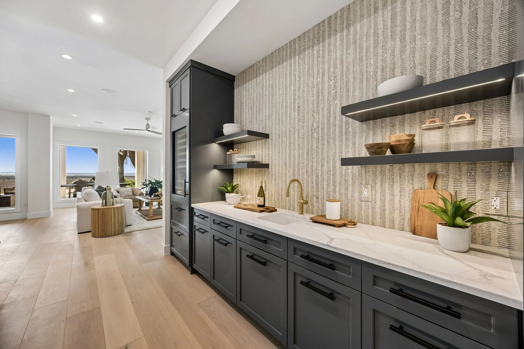 Modern kitchen with gray cabinetry, white marble countertop, open shelving with dishes and bowls, potted plants, and a textured wallpaper accent wall.