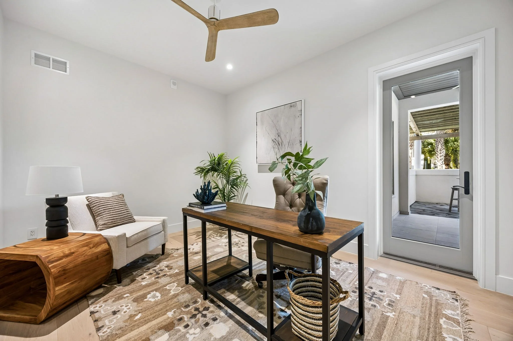 Modern room with a wooden desk, beige chair, white sofa, side table with lamp, plants, a basket, and a glass door leading outside