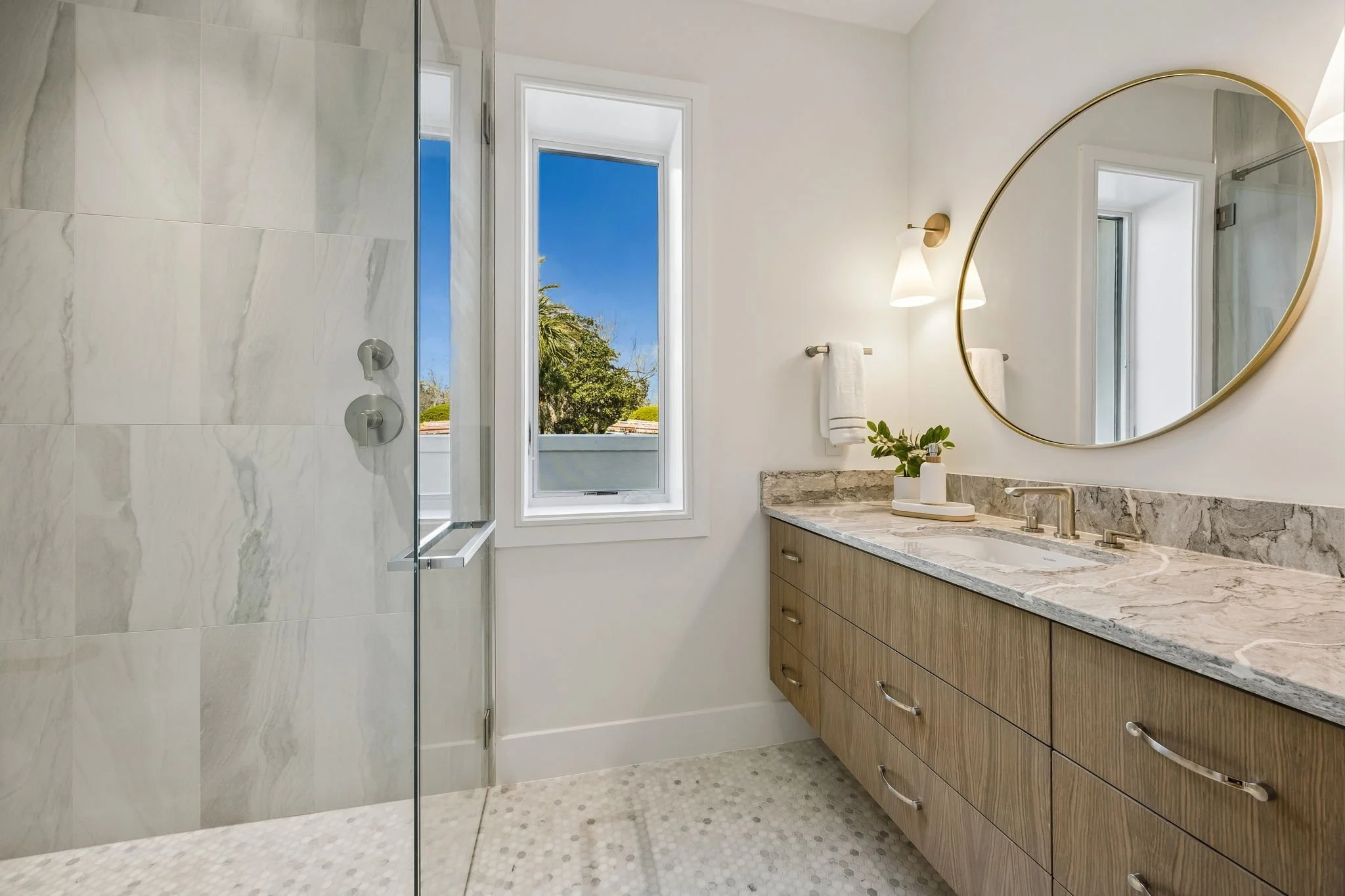 Modern bathroom with a large walk-in shower with marble tiles, a wooden vanity with a marble countertop, a round mirror, a window showing a blue sky and trees outside, and white walls.