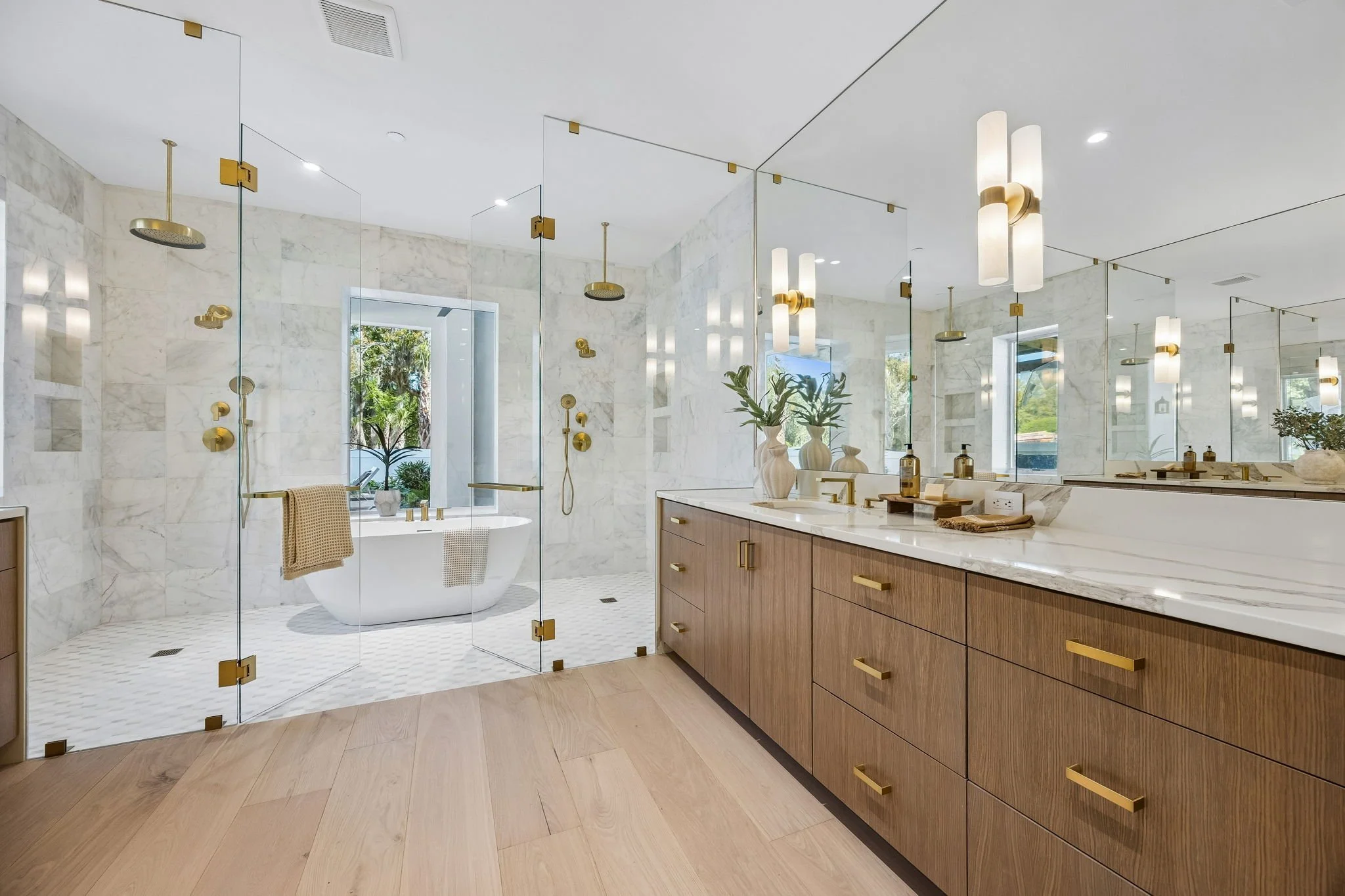 Modern bathroom with marble walls and flooring, a large mirror, wooden cabinets with gold handles, and a glass-enclosed shower area with multiple showerheads and a bathtub with a window overlooking greenery.