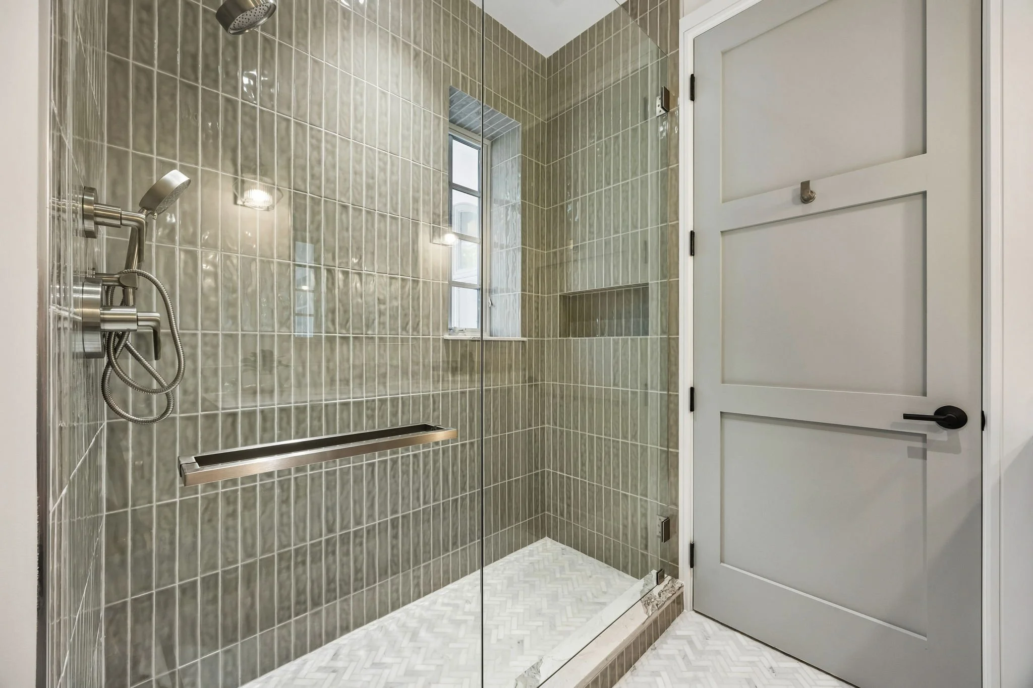 A modern walk-in shower with beige tile walls, a small window, and a glass door. The shower has a chrome showerhead, a handheld shower wand, and built-in shelves. The floor has a herringbone pattern, and there is a white door outside the shower.