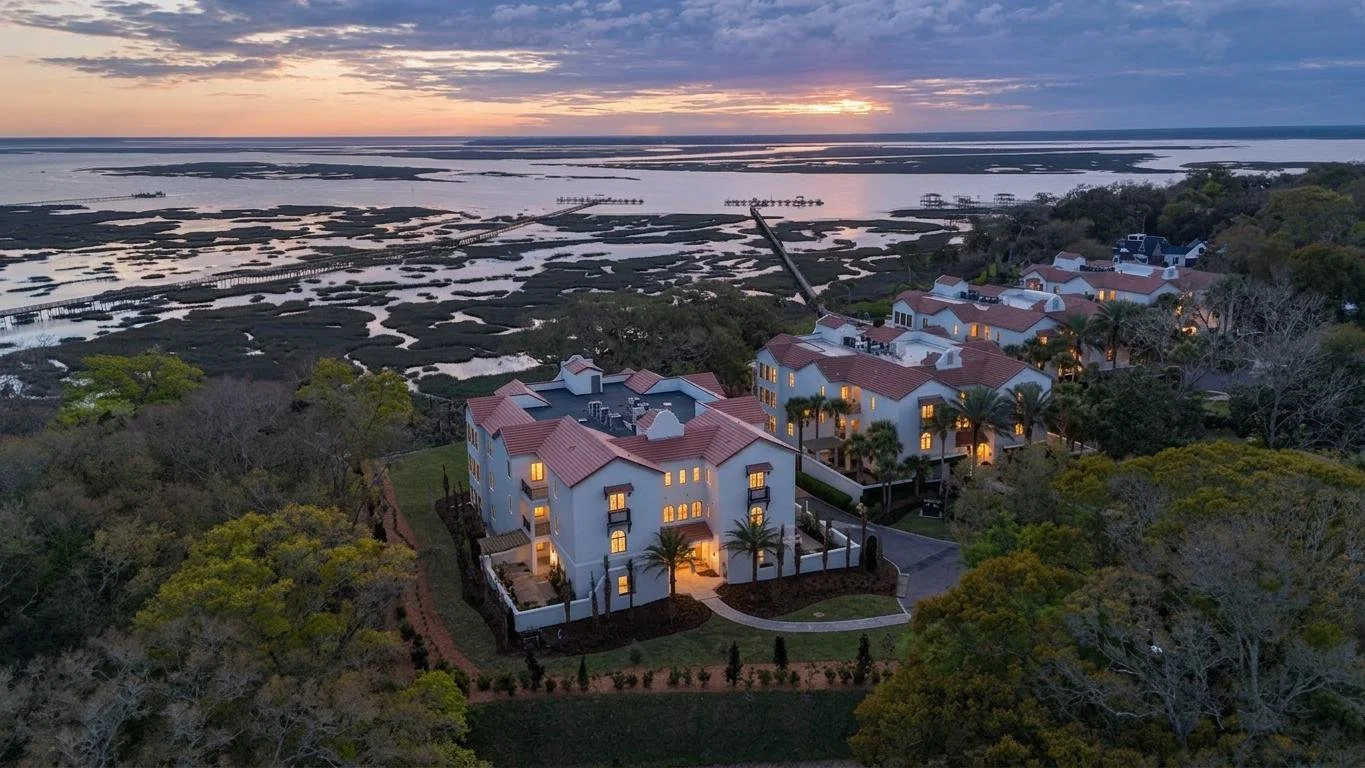 Aerial view of a coastal residential area at sunset with multiple white houses with red tile roofs surrounded by greenery, water, and wetlands.