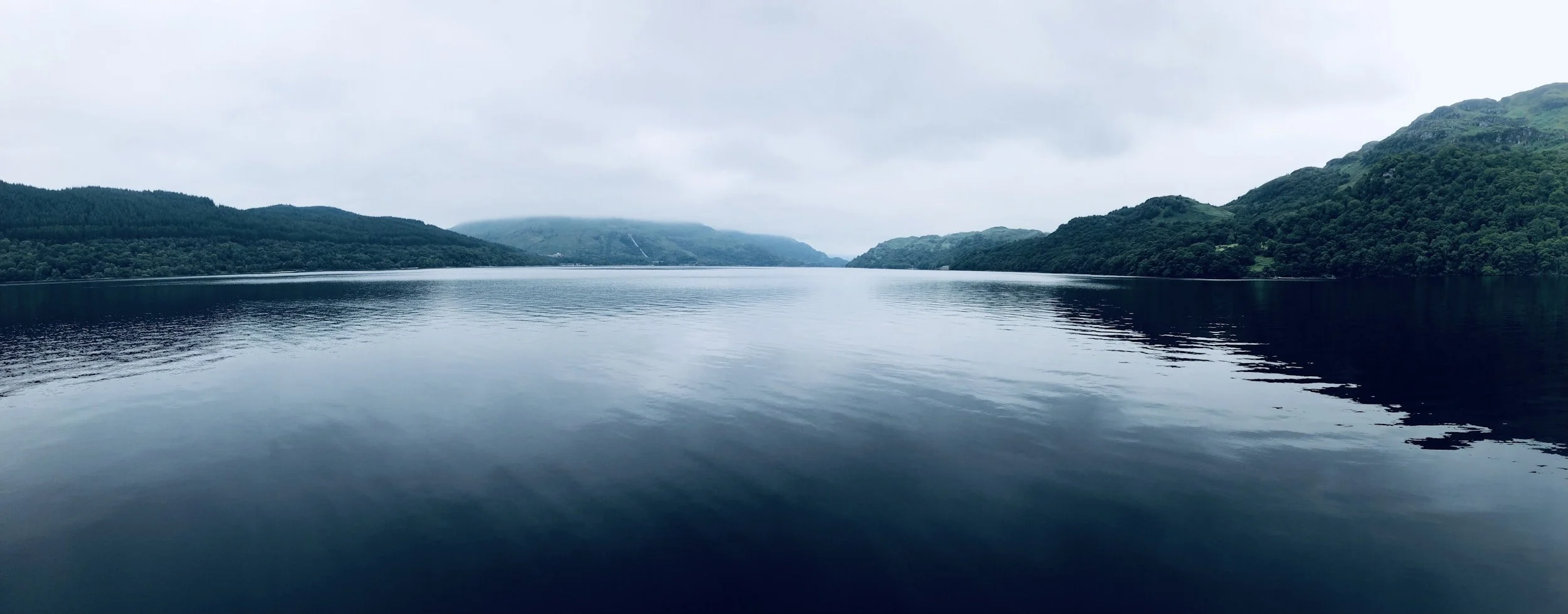A calm lake surrounded by green hills and mountains under a cloudy sky in Scotland.