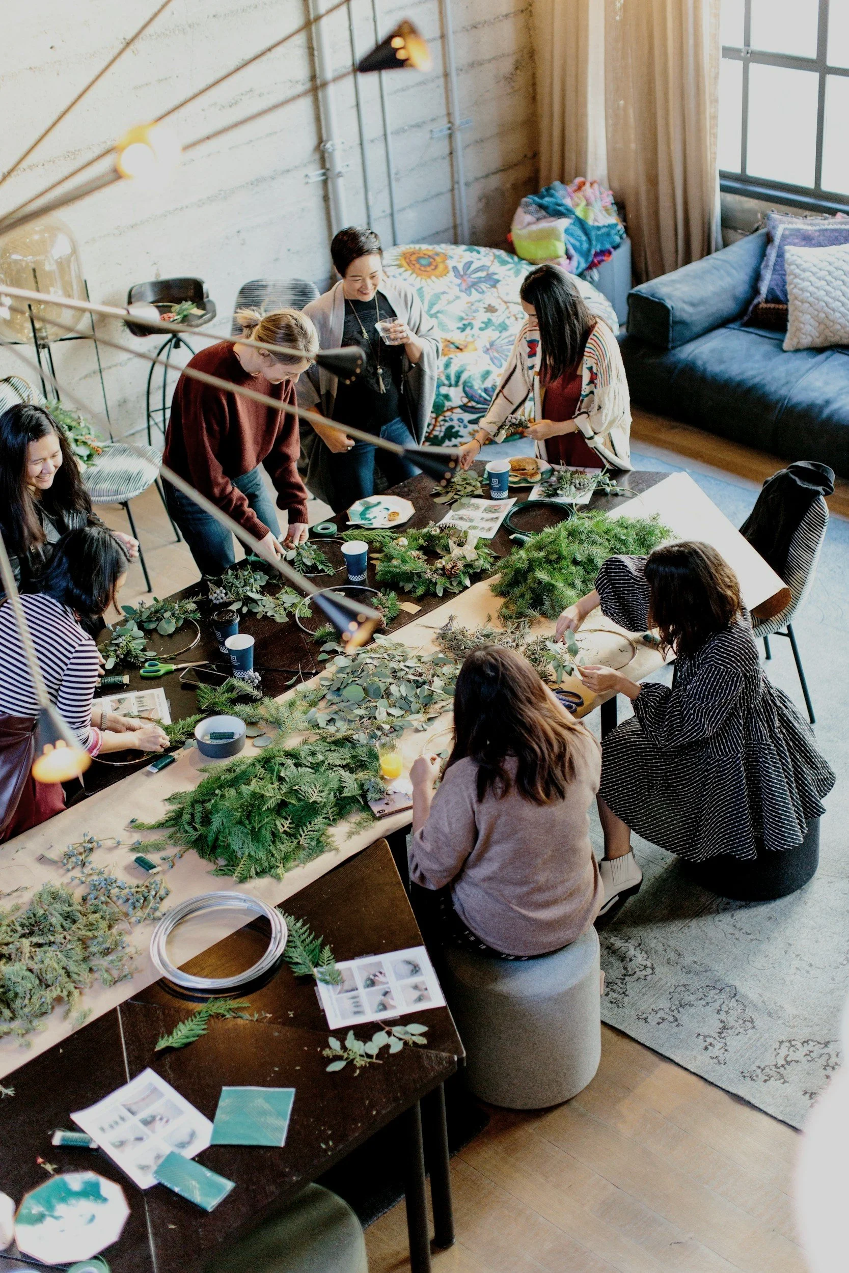Group of women making holiday wreaths around a large wooden table in a cozy indoor space with natural light, plants, and colorful decor.