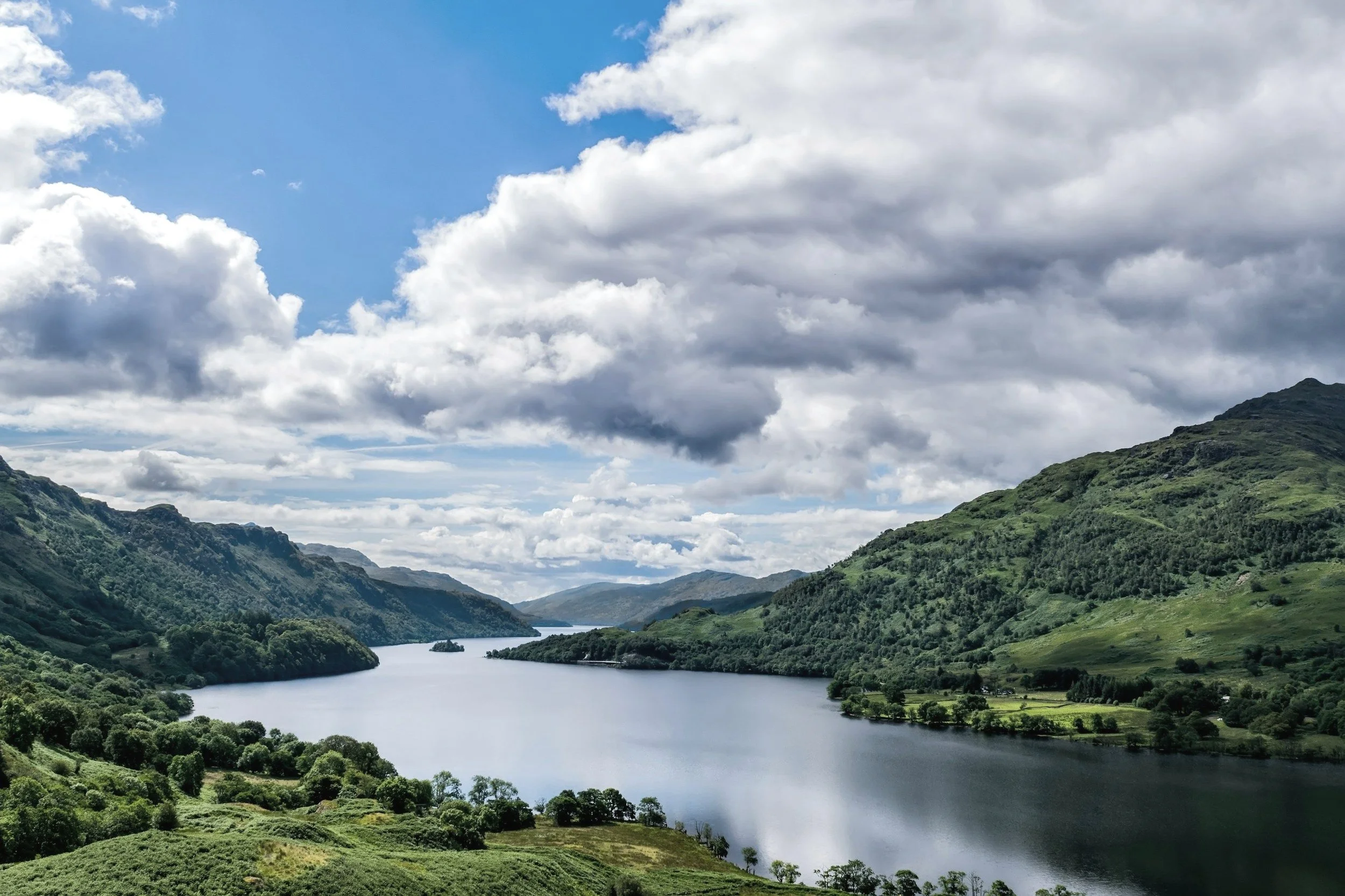 Scenic view of a river flowing through lush green mountains under a partly cloudy sky in Scotland.