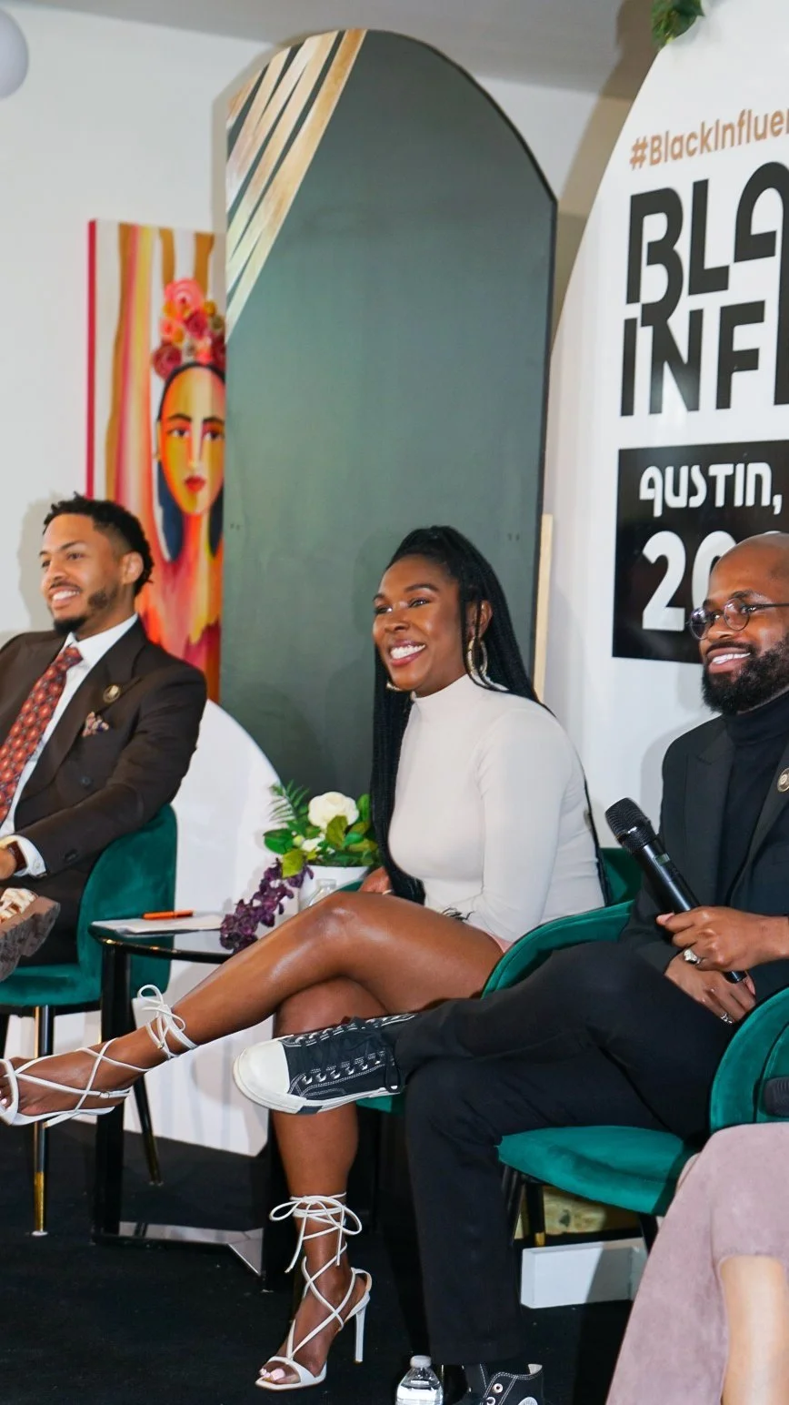 Three people sitting on green chairs at a panel discussion, smiling, with a woman in a white dress, men in suits, and a microphone.