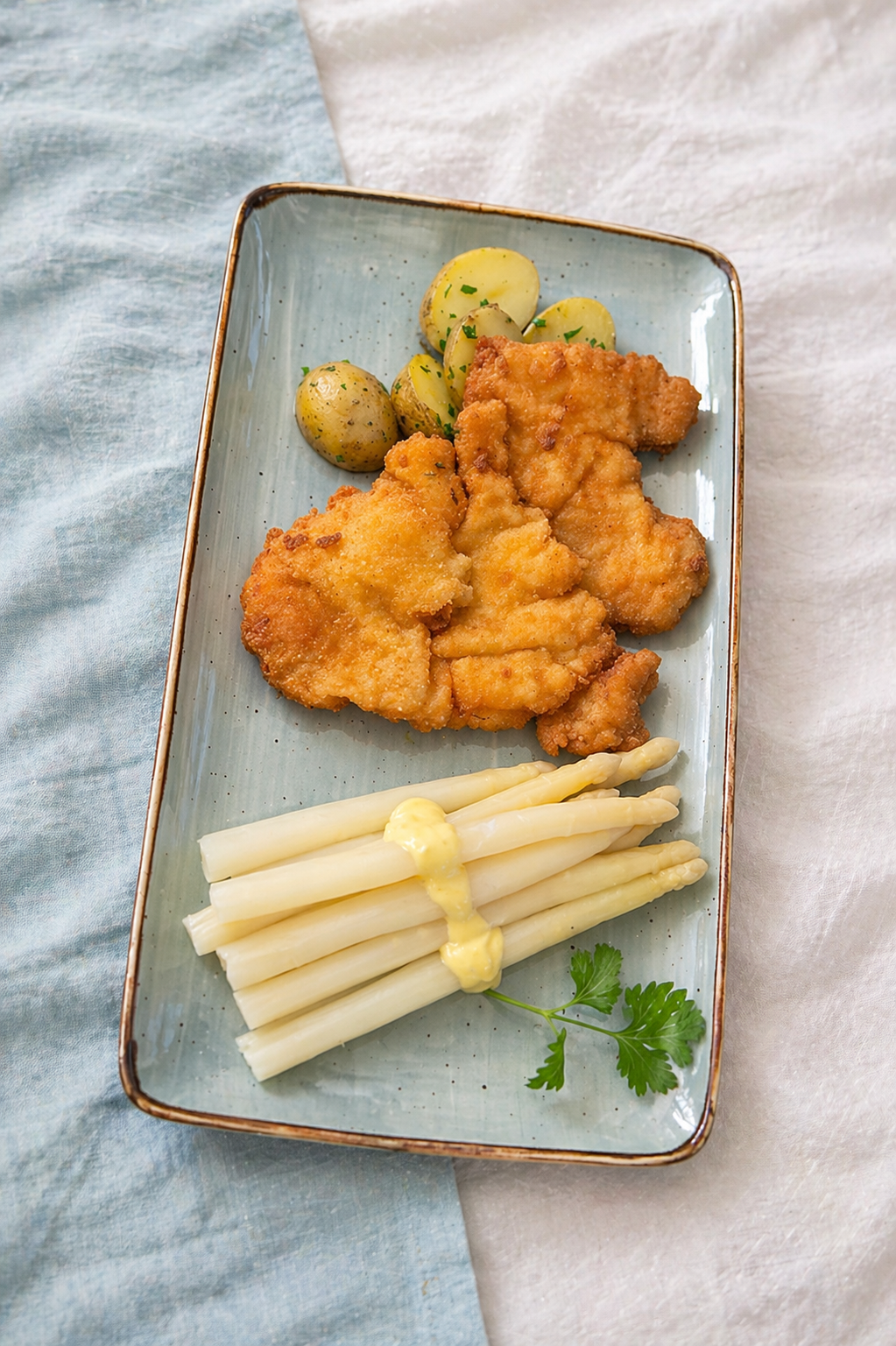 Gefülltes Hähnchenfilet mit weißen Spargeln, Kartoffeln und Petersilie auf einem blauen rechteckigen Teller.