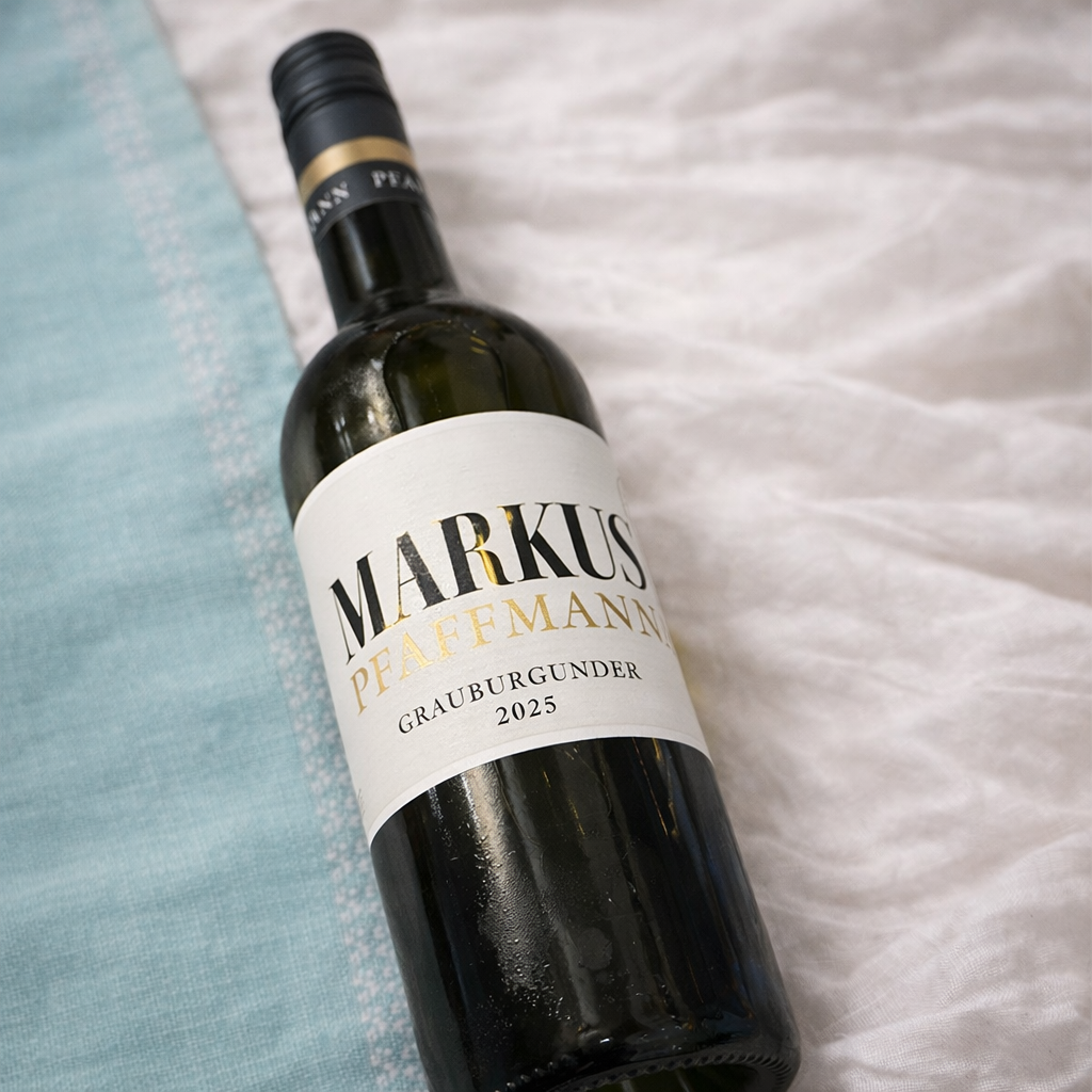Eine Flasche Wein mit einem weißen Etikett, das den Namen 'Markus Pfaffmann' und die Bezeichnung 'Grauburgunder 2025' trägt, liegt auf einer weißen Stoffoberfläche neben einem blauen Stoff.
