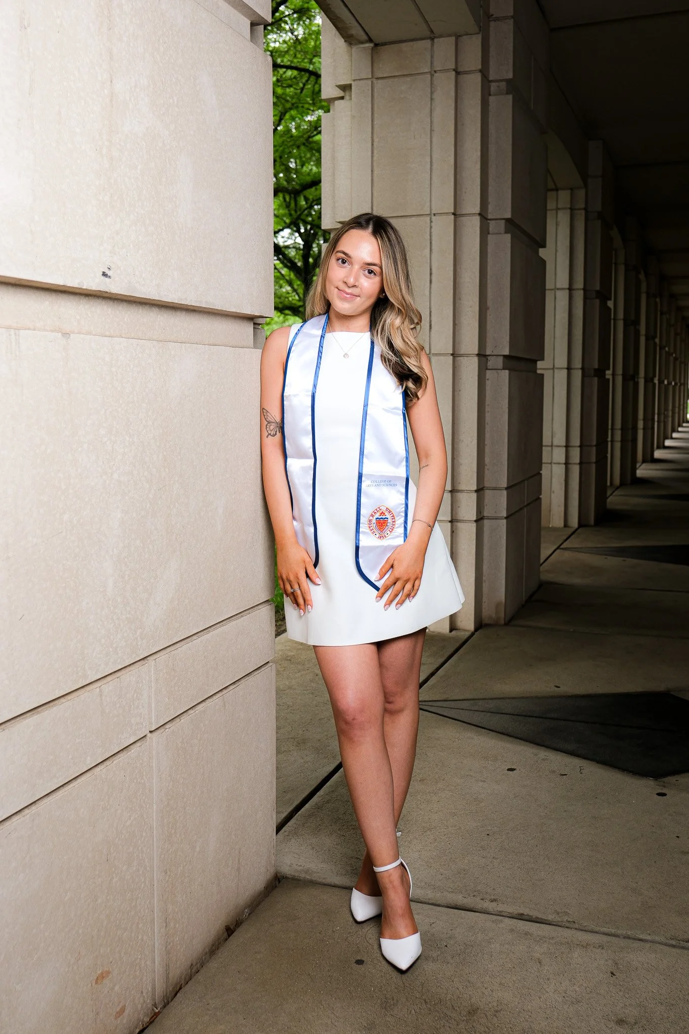A young woman is wearing a white college graduation dress with blue trim and a graduation stole, standing outdoors on a stone sidewalk near a large stone building with columns, smiling at the camera.