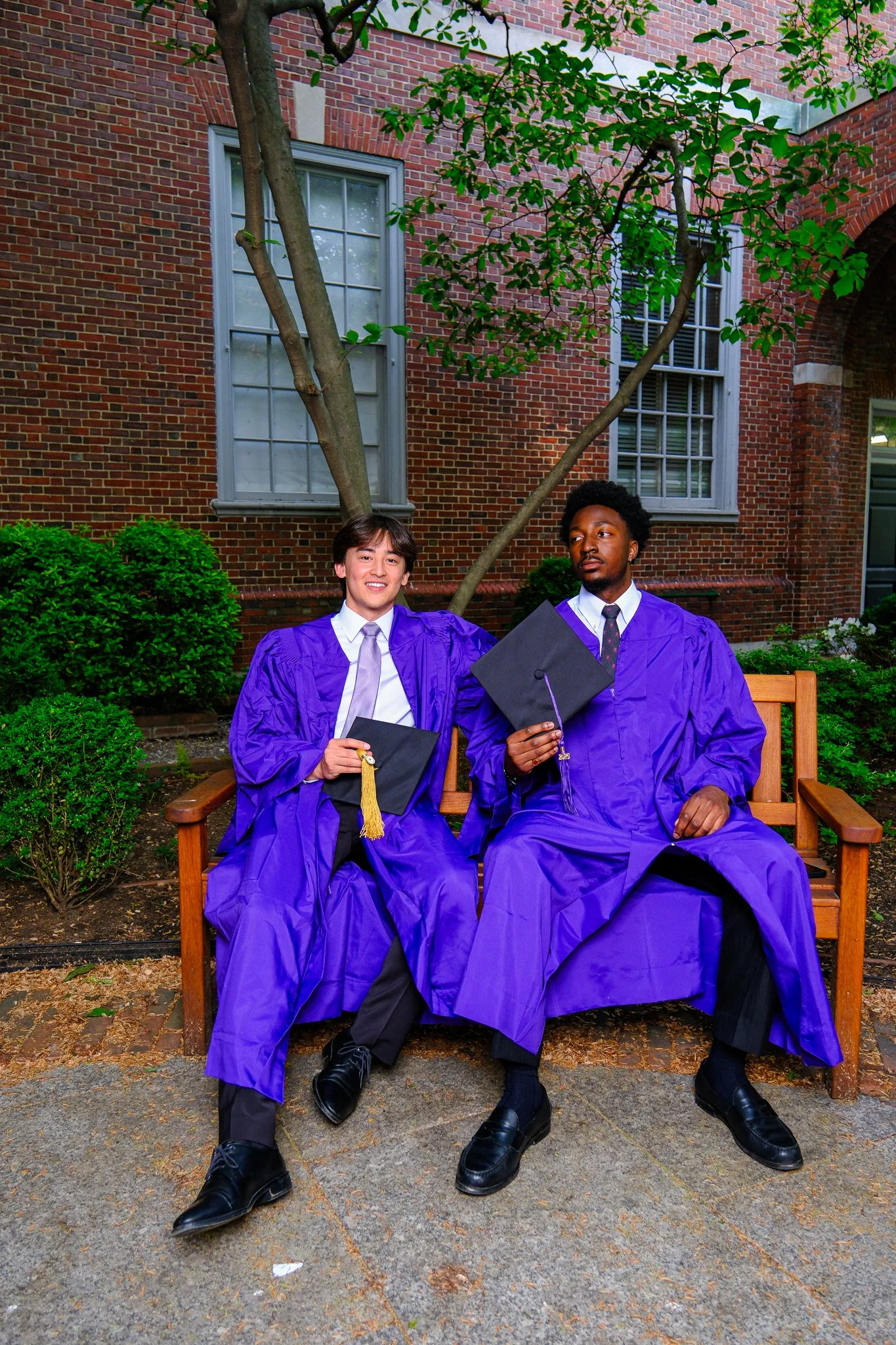 Two graduates in purple gowns and caps sitting on a wooden bench outdoors in front of a brick building with trees and bushes, celebrating graduation.