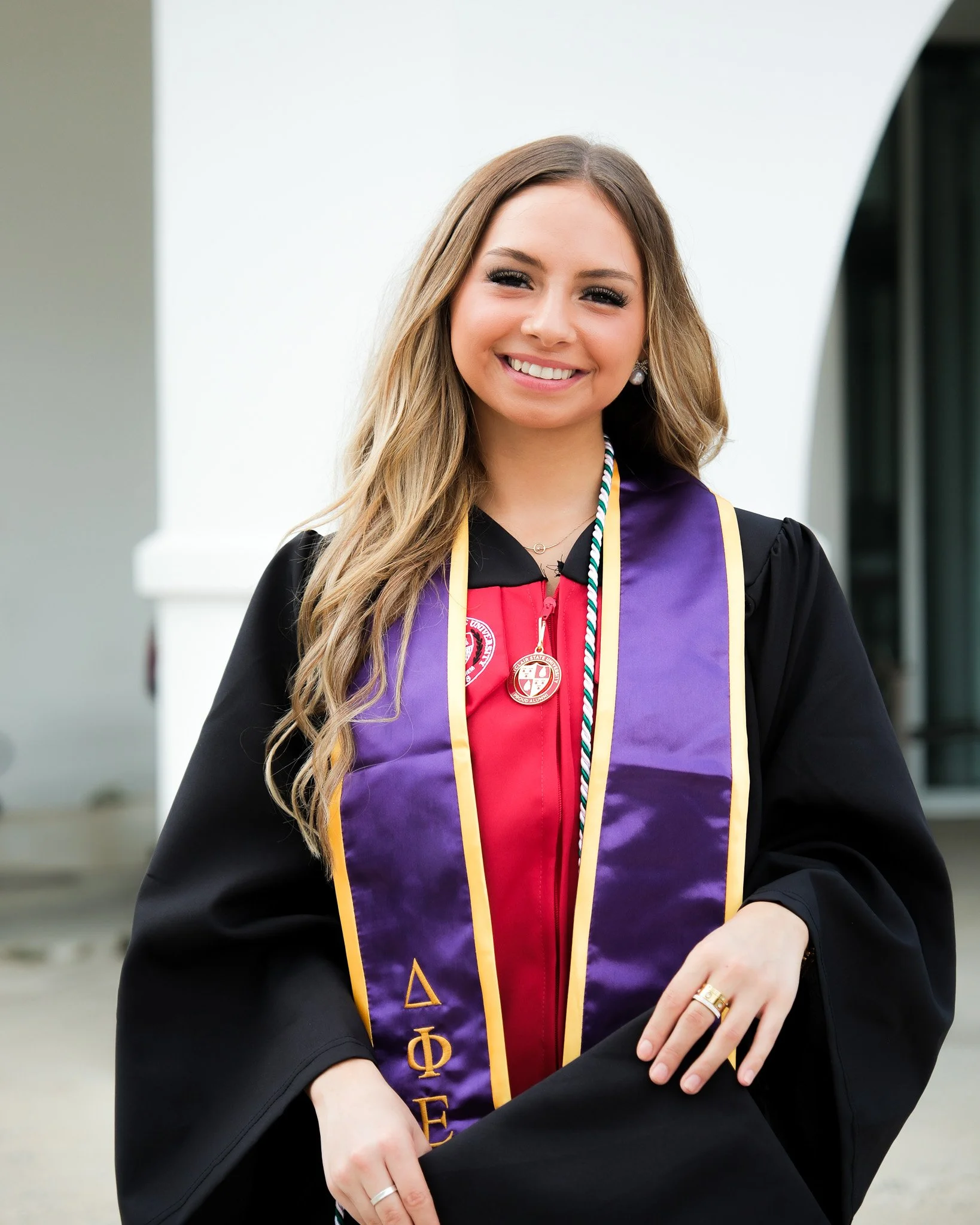 Young woman in graduation gown and honor cords, smiling outside.