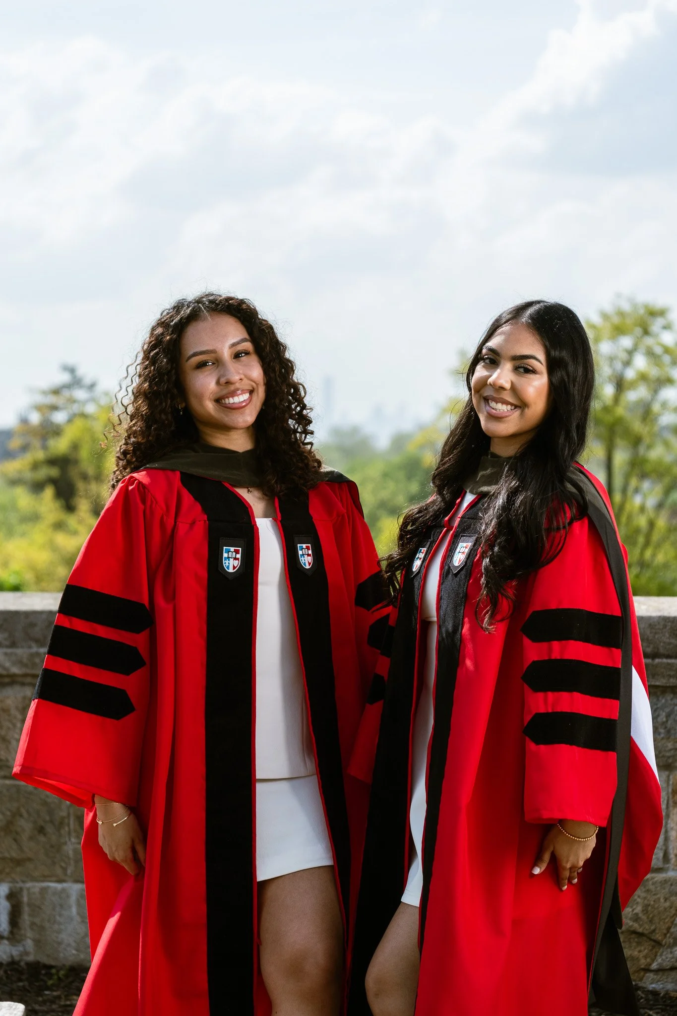 Two women in graduation caps and gowns standing outdoors, smiling at the camera, with trees and a city skyline in the background.