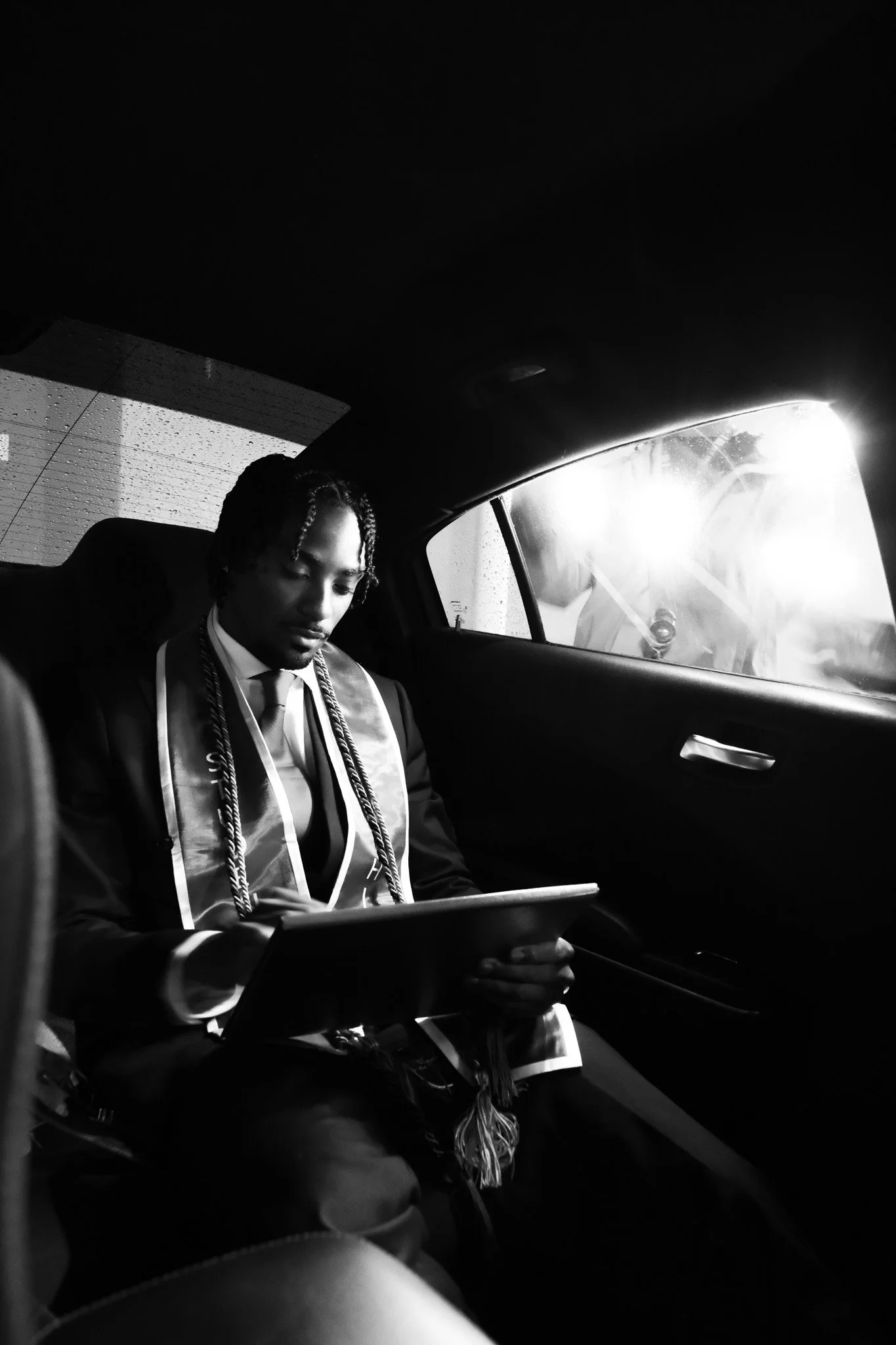 A man in formal attire sitting in the backseat of a car, reading a book, with sunlight streaming through the window. It appears to be a rainy day outside.
