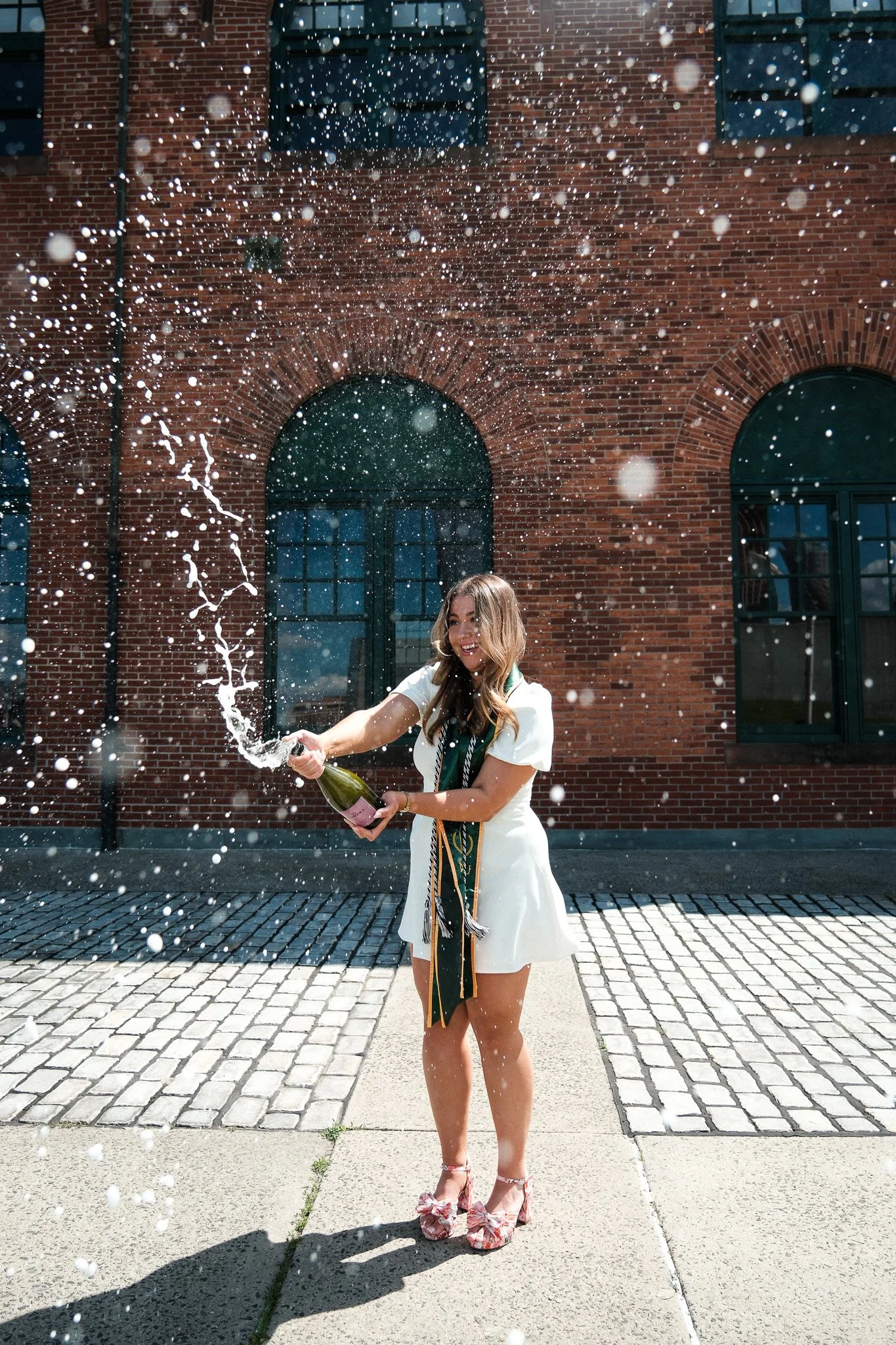 A woman celebrating outdoors in front of a brick building, opening a bottle of champagne that is spraying.