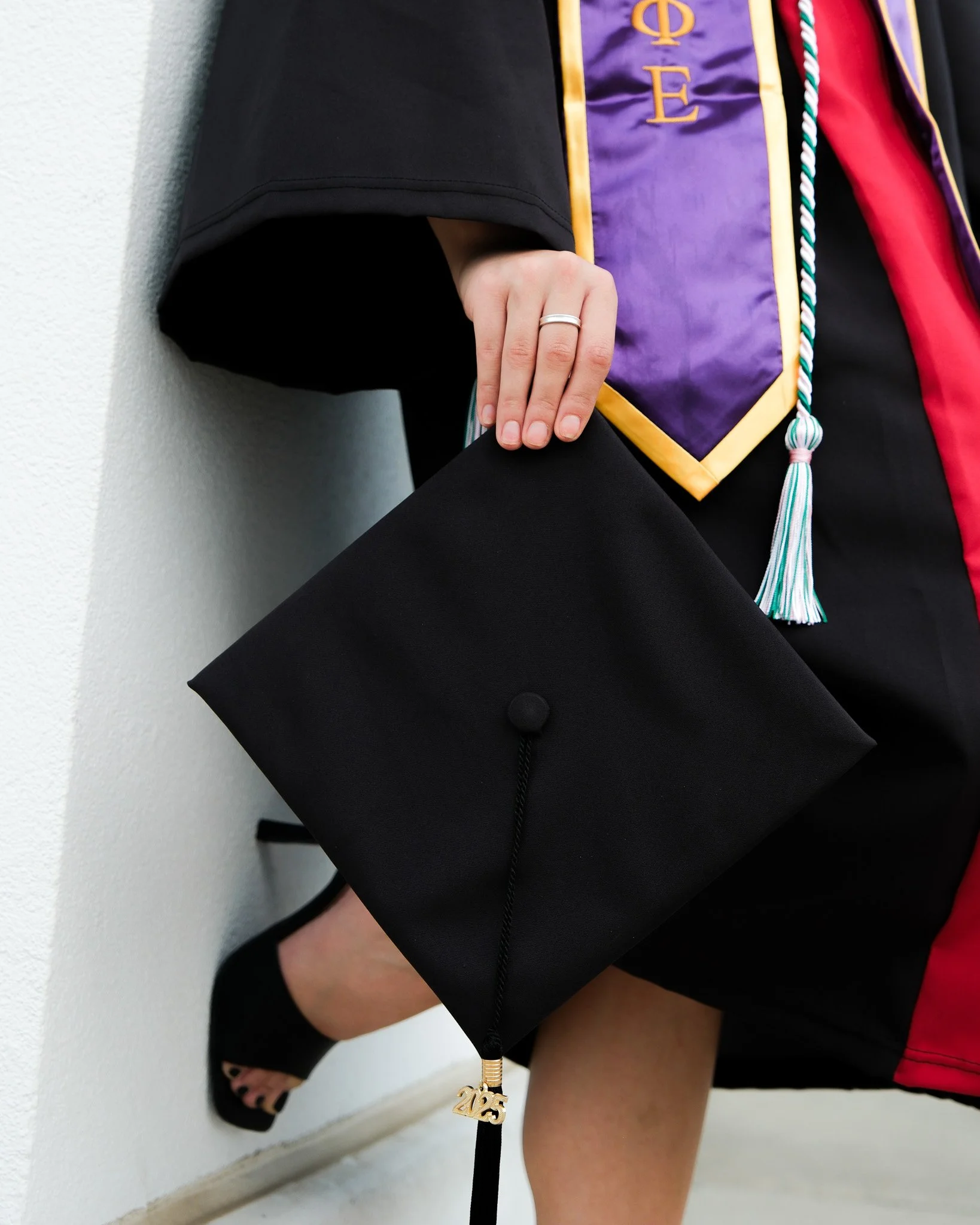 A person holding a black graduation cap with a gold '2025' charm. The individual is dressed in graduation attire, including a black gown, and is standing against a white wall.