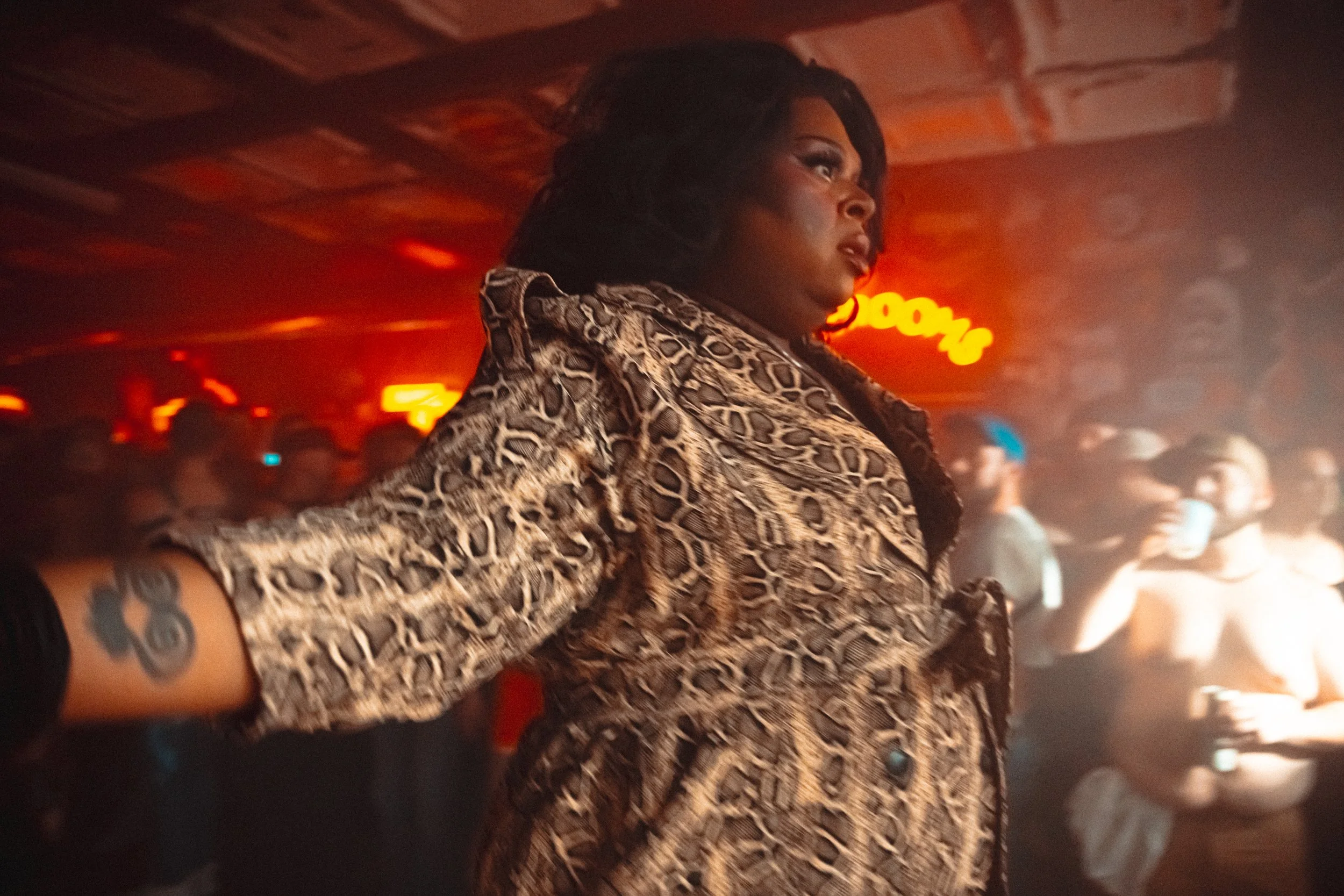 A woman with dark hair and dark skin wearing a leopard print jacket with her arm outstretched at a nightclub or concert. The background shows a crowd of people and a red neon sign.