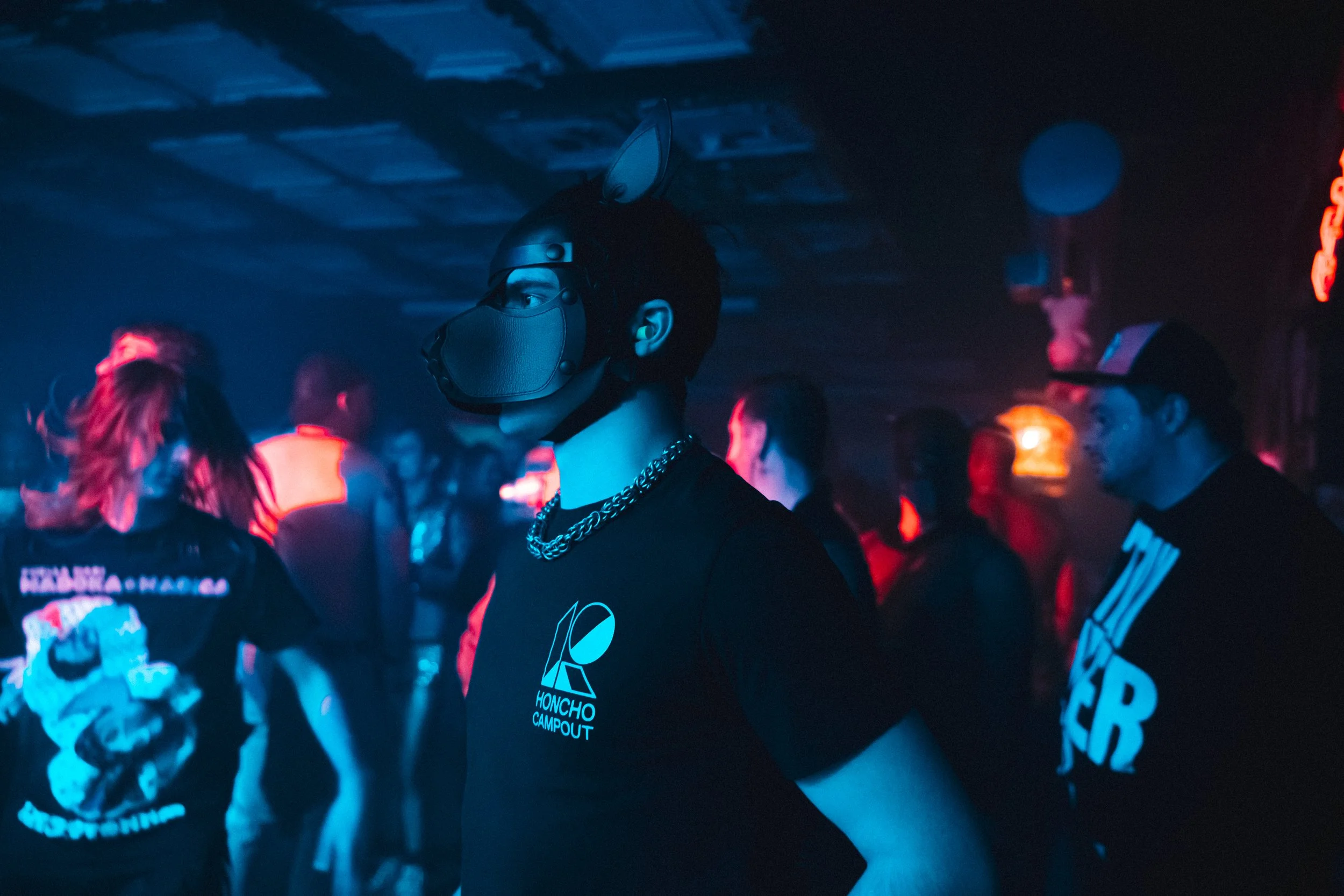 Person wearing a black horse mask and black T-shirt with 'Honcho Campout' logo in a dark, neon-lit club with other partygoers.