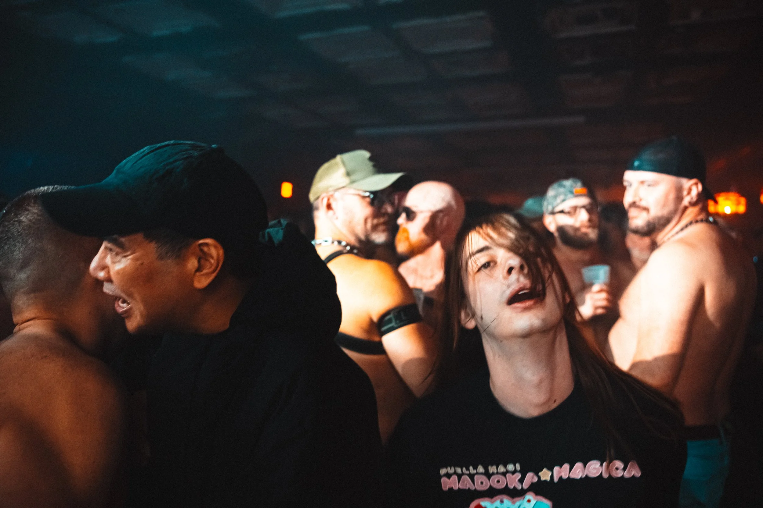 A group of people at a party or concert, some shirtless and wearing hats, with one woman in the foreground with wet hair and an expressive face, in a dimly lit environment.
