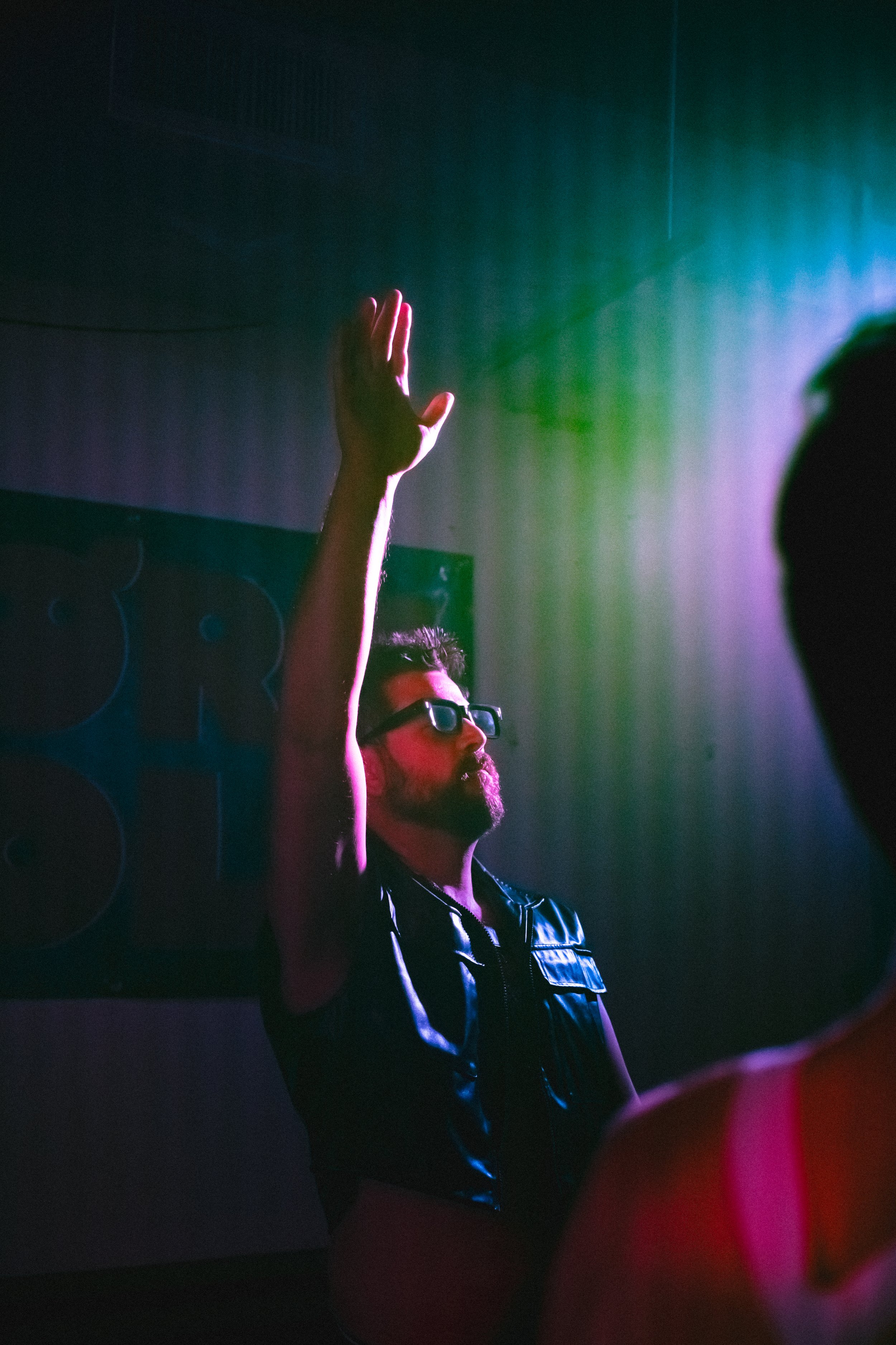 A bearded man with glasses in a leather vest raising his hand at a party or club, with colorful lighting creating a vibrant atmosphere.