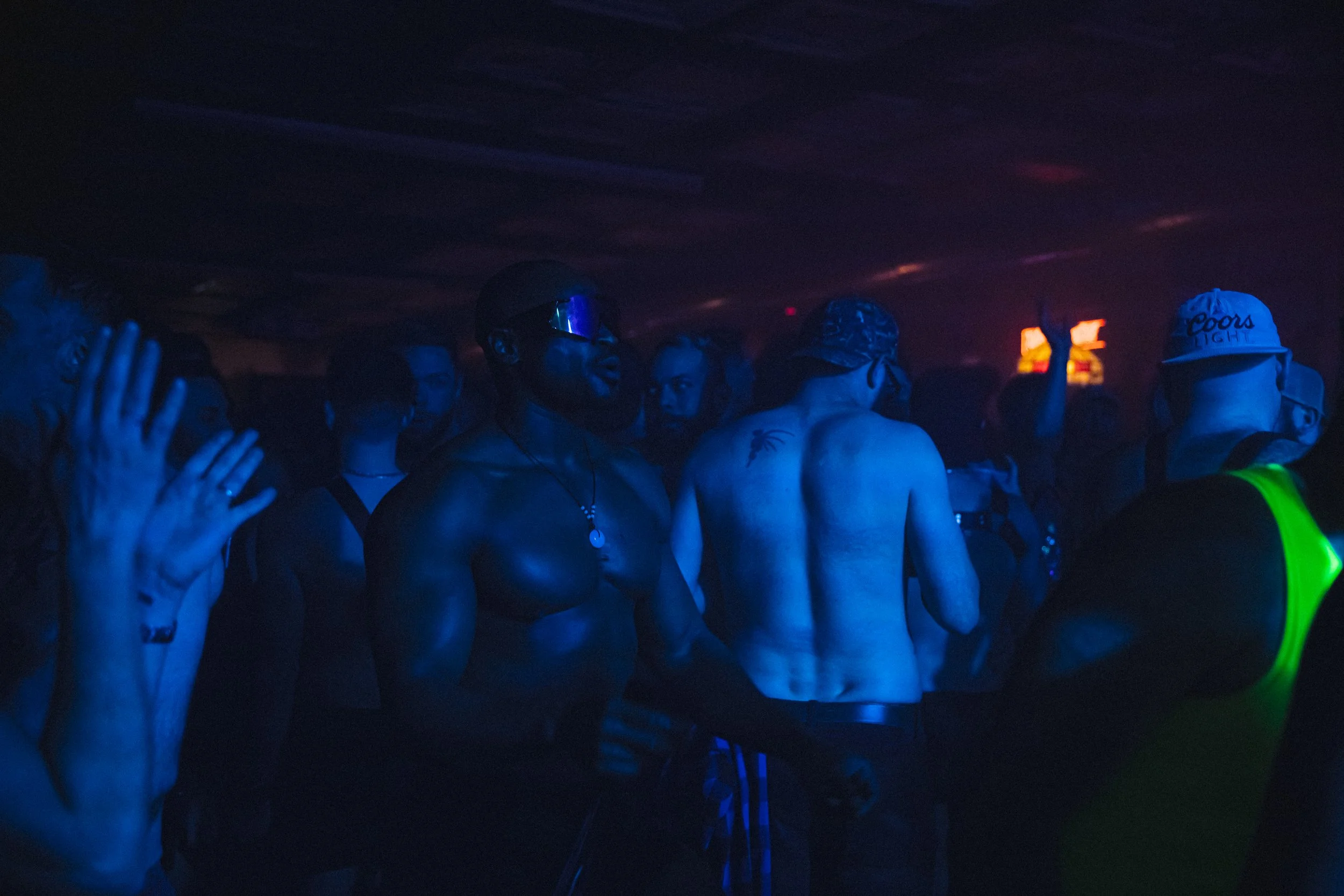 People dancing and enjoying themselves at a dark, neon-lit nightclub or party, some with sunglasses and minimal clothing, with one person raising their hands.
