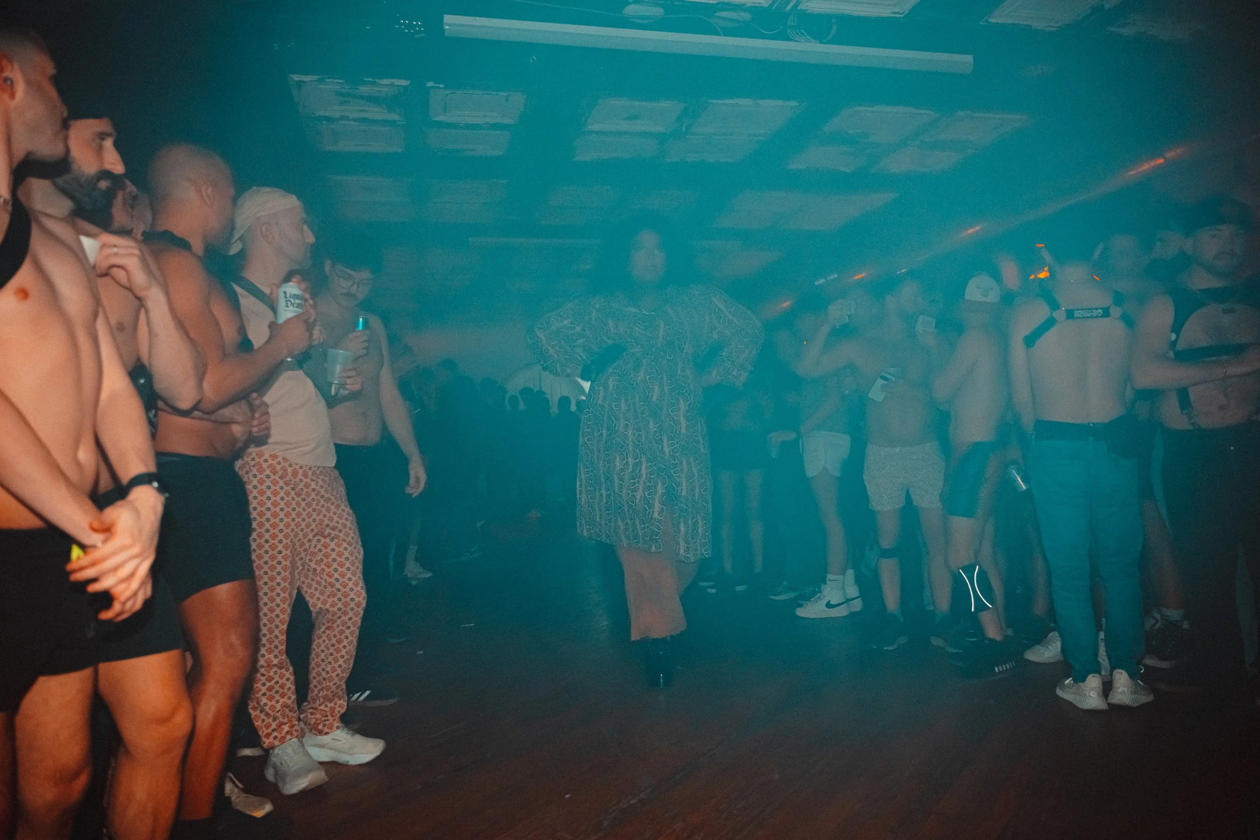 Person dressed in a patterned dress standing in the center of a crowded room with people. The room has a dark ceiling with square panels, and a blue smoke or fog effect is present.