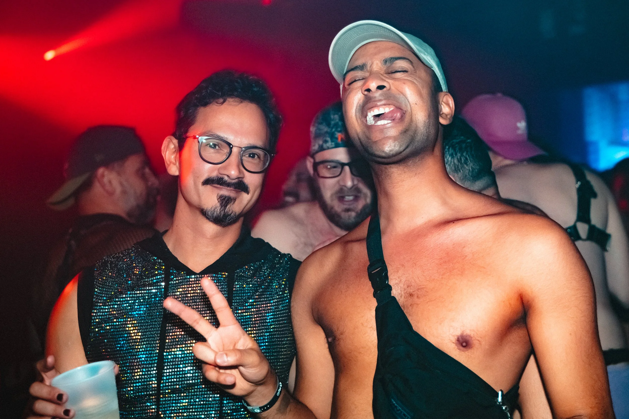 Two men enjoying a party with colorful lighting, one making a peace sign and the other shirtless with a bag strap across his shoulder.