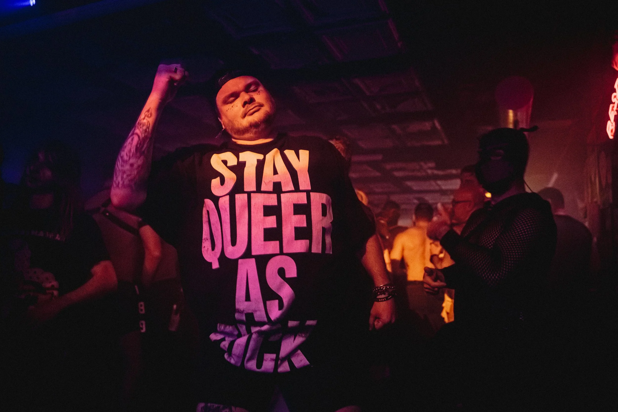 A person at a nightclub or concert venue wearing a black T-shirt that says "STAY QUEER AS" in bold letters. The individual has tattoos on their arm, jewelry, and a black cap, with a serious or contemplative expression. In the background, other people