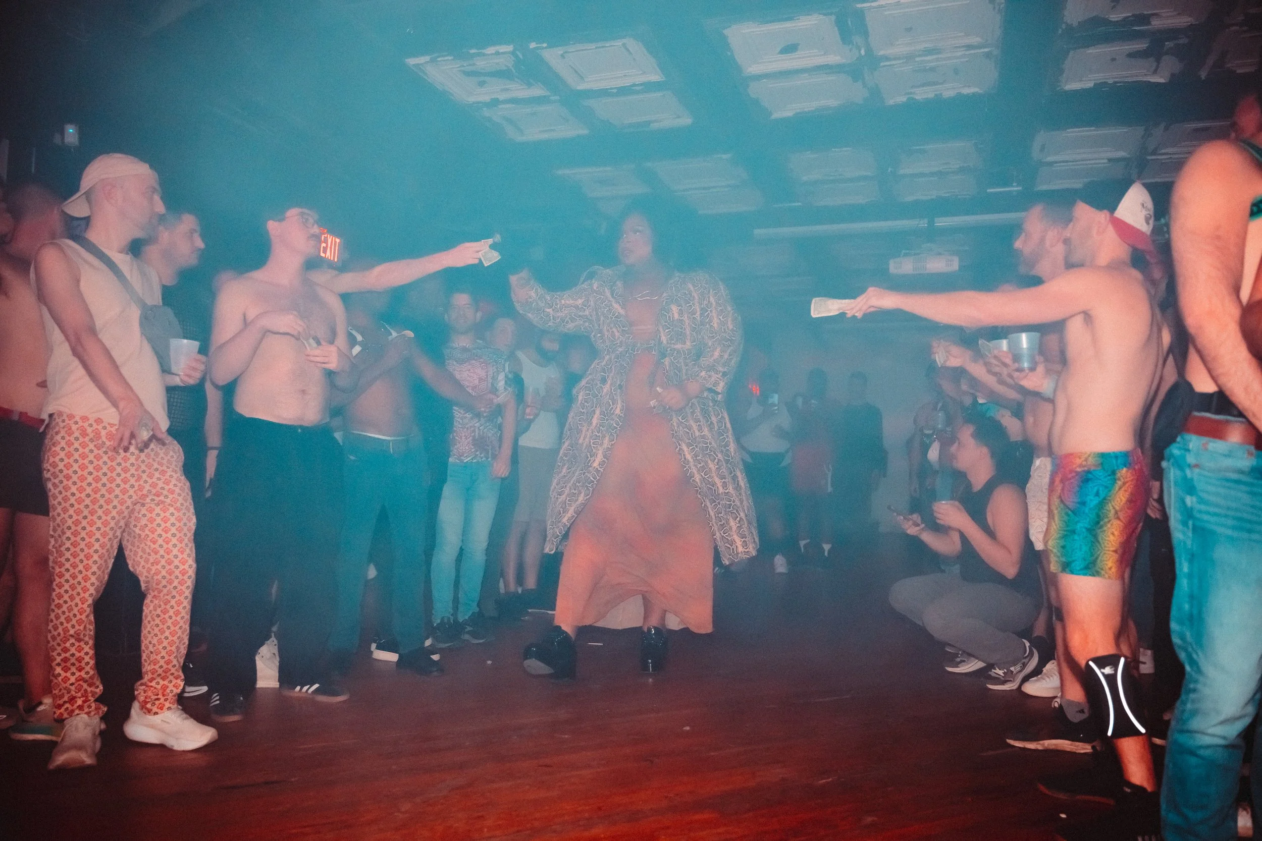 A drag performer stands confidently in the center of a dance floor, dressed in an orange gown and a patterned coat, surrounded by an audience of men in casual, summer attire, some shirtless, reaching out towards the performer. The setting is a dimly 