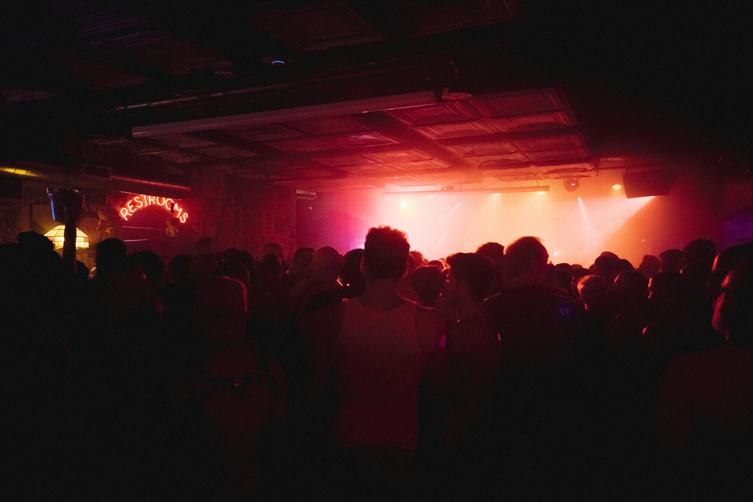 Crowd of people watching a concert or performance on stage in a dark venue with pink and orange lighting.