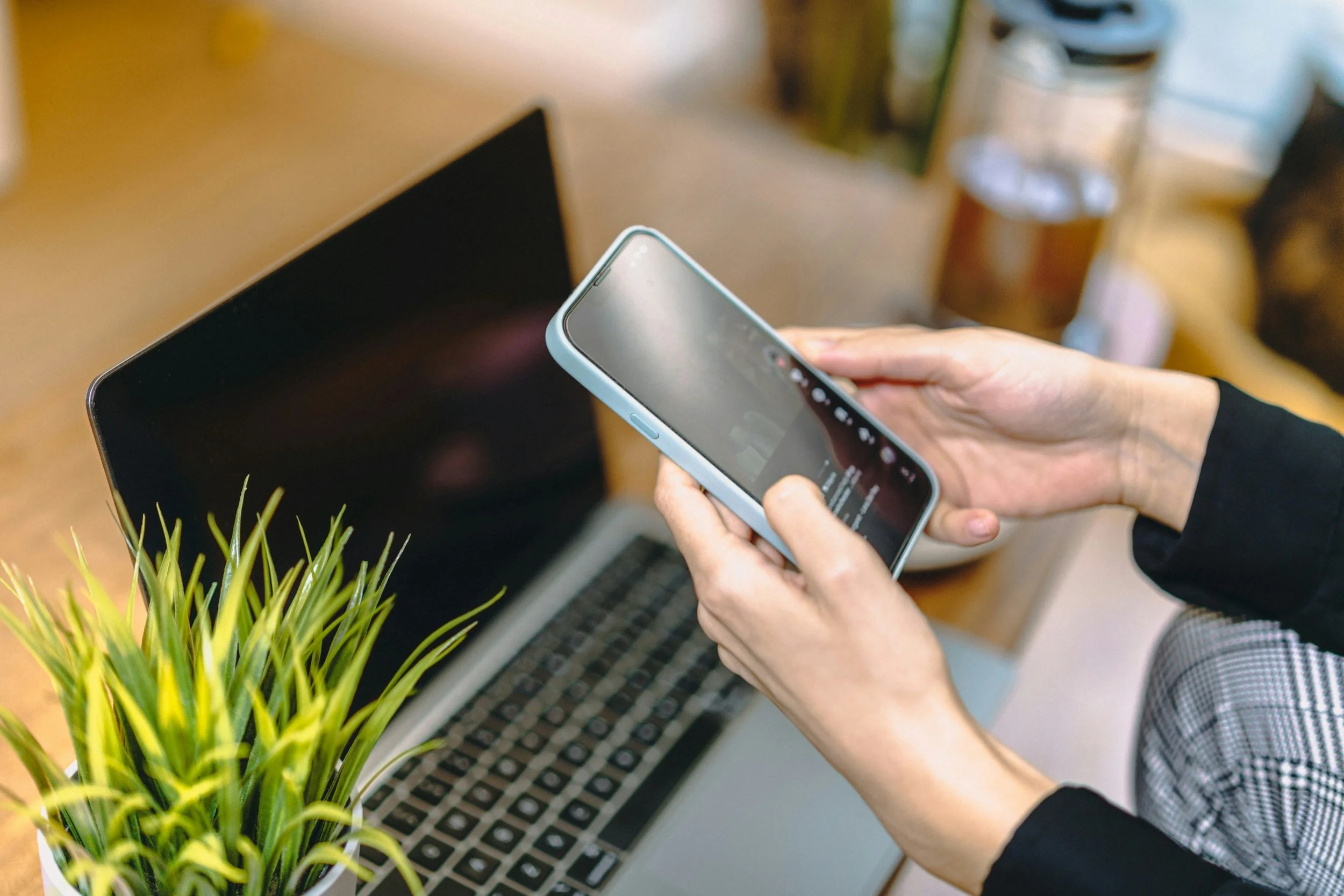 Two hands holding a cellphone with a laptop in the background