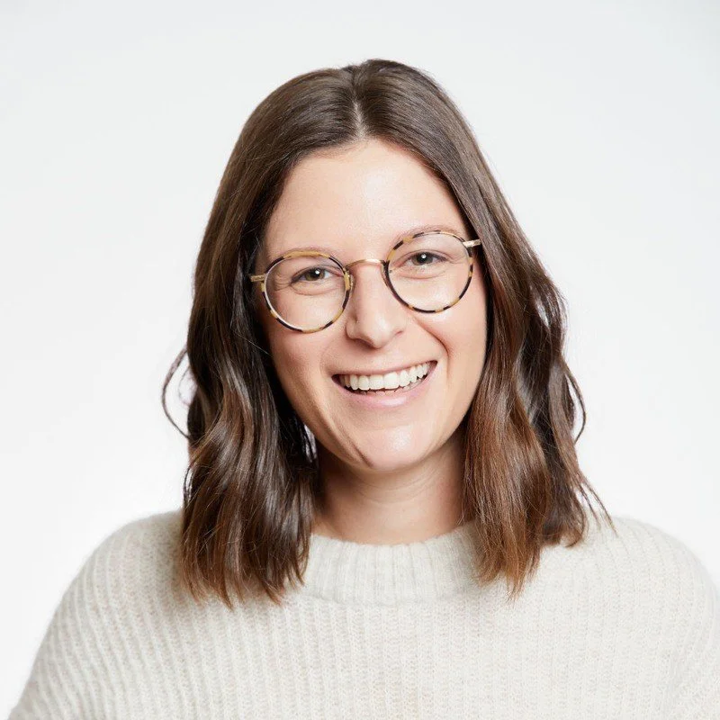 A woman with shoulder-length brown hair, wearing glasses with round frames and a white knit sweater, smiling against a plain white background.