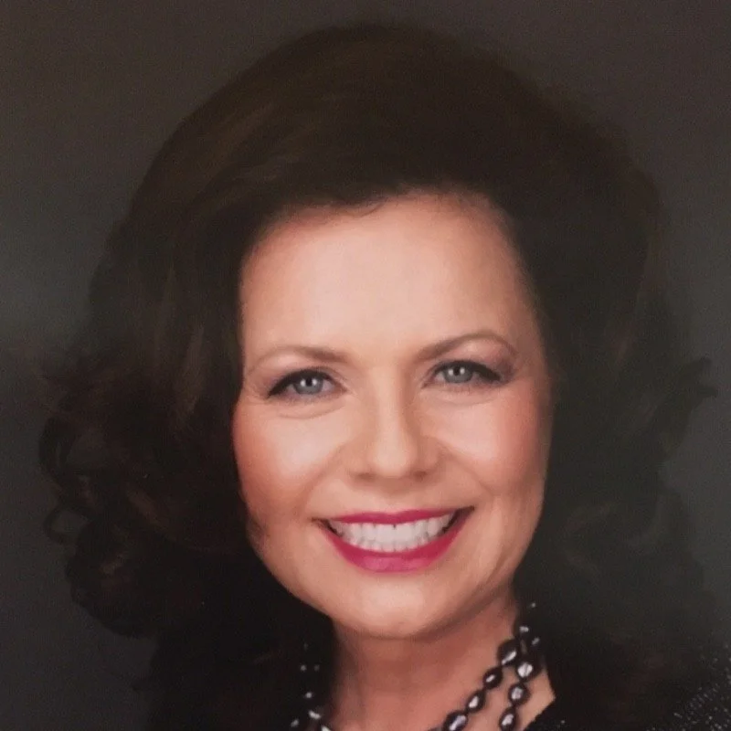 Close-up portrait of a smiling woman with dark, curly hair and light makeup, wearing a black top and a pearl necklace.