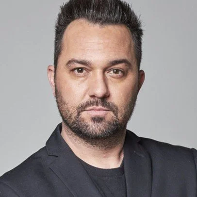 Portrait of a man with dark hair and a beard, wearing a black blazer and shirt, against a gray background.