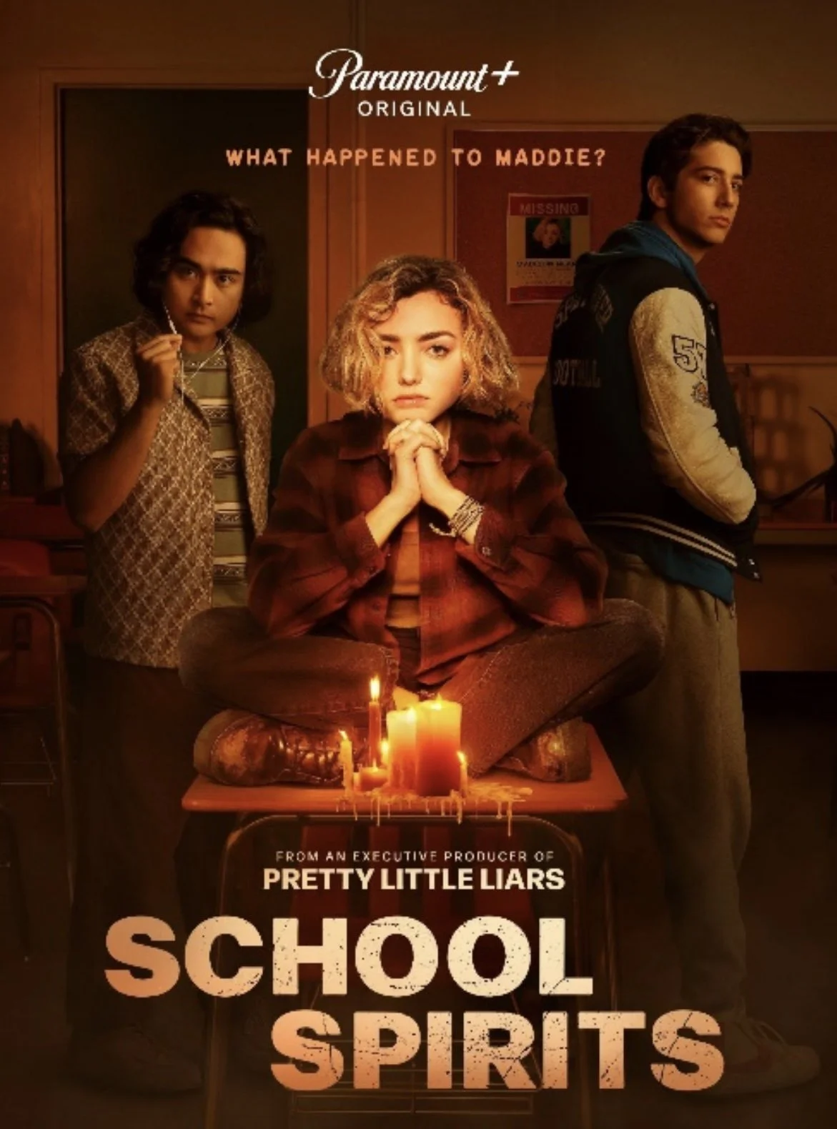 Poster for the series 'School Spirits' with three teenagers, one sitting cross-legged on a desk with candles, and two standing on either side, in a dimly lit classroom.