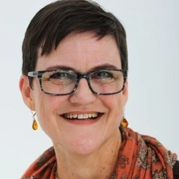 A woman with short dark hair, glasses, and a patterned scarf smiling in front of a light background.