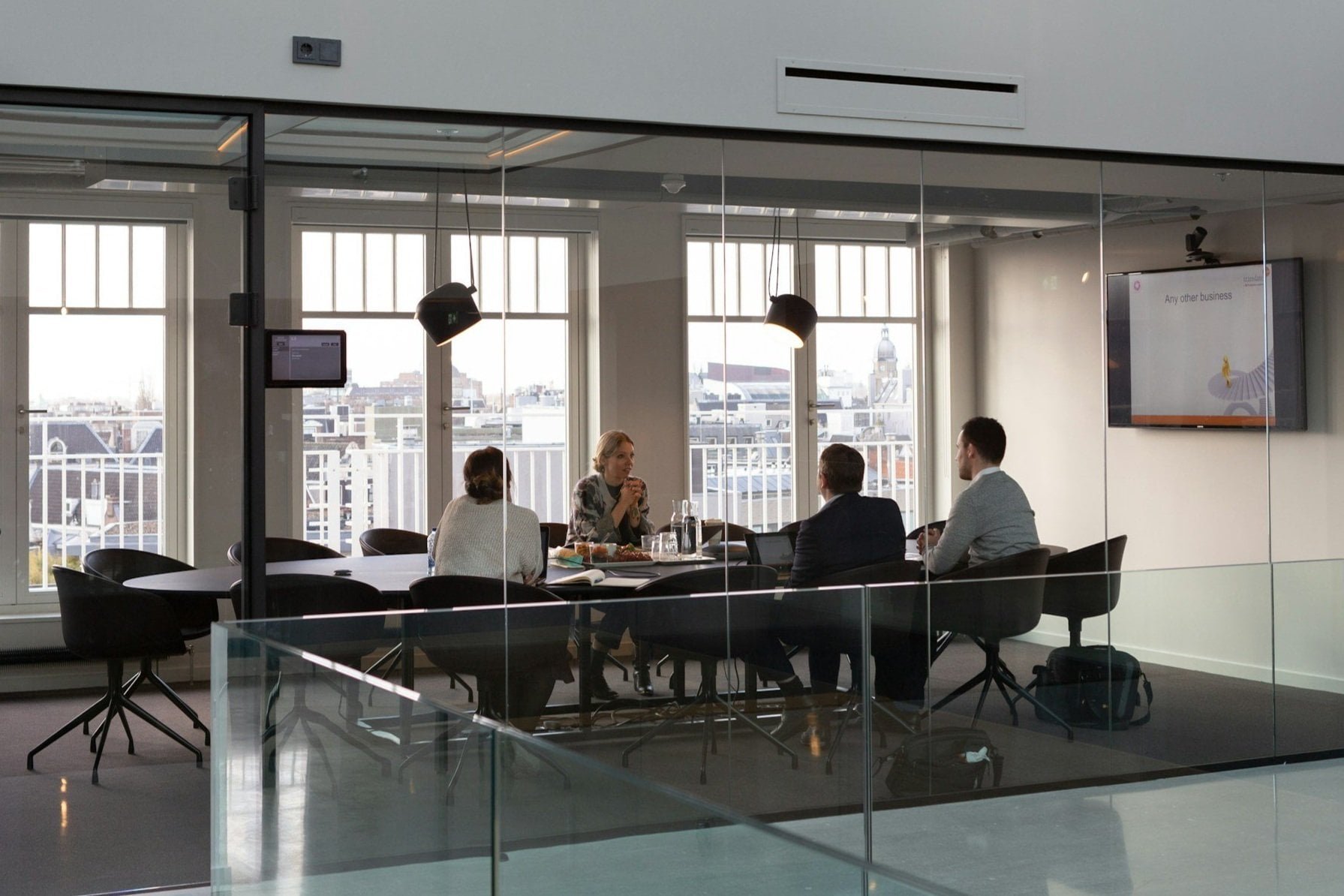 Four business people in a meeting room with large windows and city view, three facing the woman speaking, sitting at a round table with laptops, water bottles, and documents.
