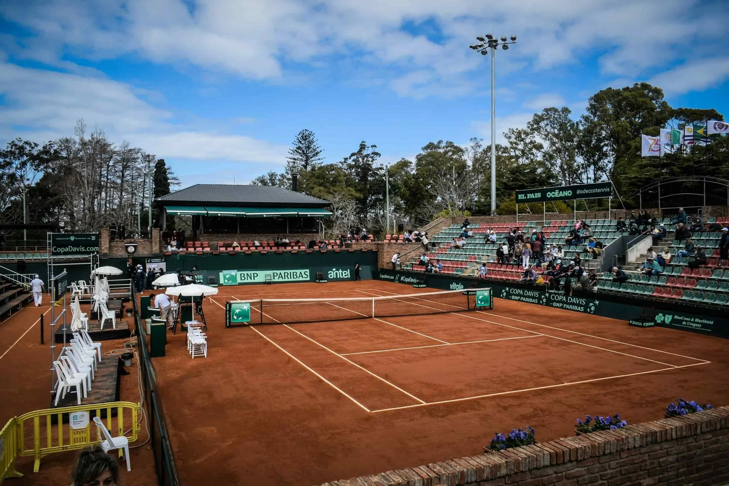 Empty outdoor clay tennis court with seating stands, banners, and a small pavilion under partly cloudy sky at a professional tennis tournament.