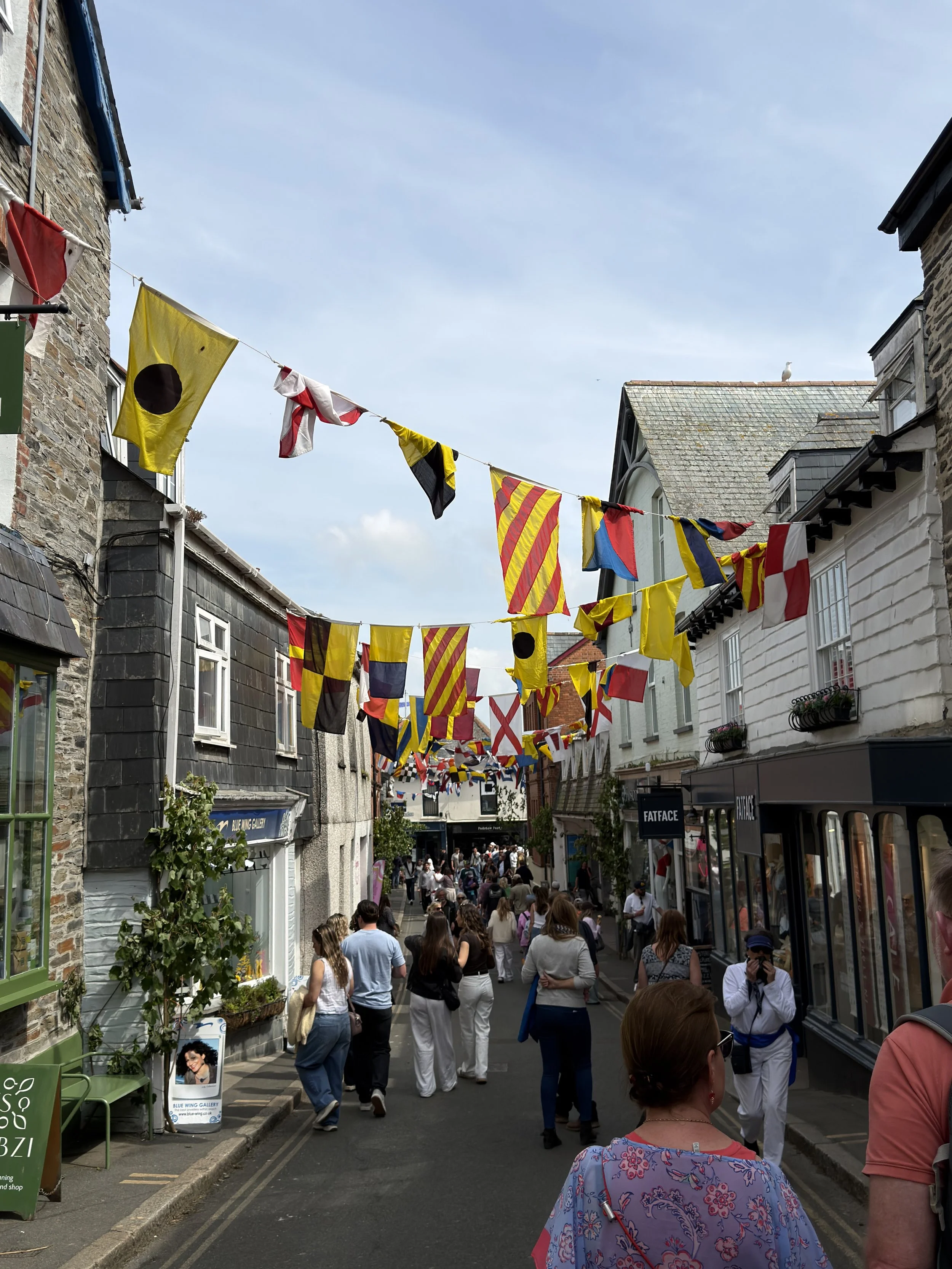Crowd of people walking down a lively street decorated with colorful flags hanging overhead. Shops with storefronts and buildings with different styles line both sides of the street under a clear blue sky.