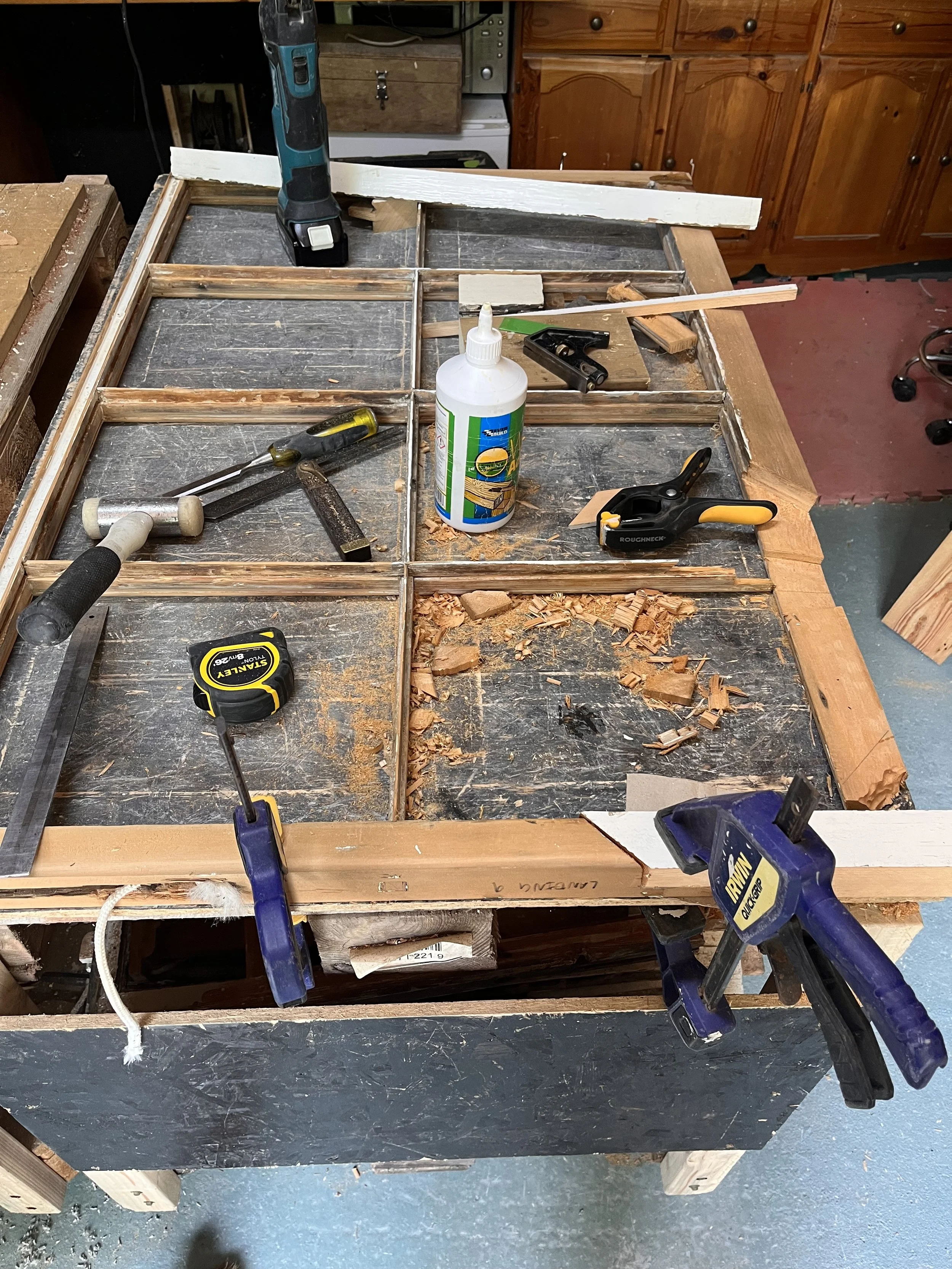 A woodworking workbench with various tools including clamps, a hammer, a hand saw, a square, a glue bottle, and a cordless drill, surrounded by wood shavings and scraps in a workshop.